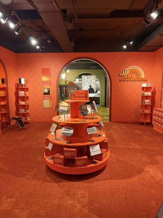 Retro orange-red boutique interior with a circular tiered display of boxed goods centered on carpet, arched doorway revealing green room.
