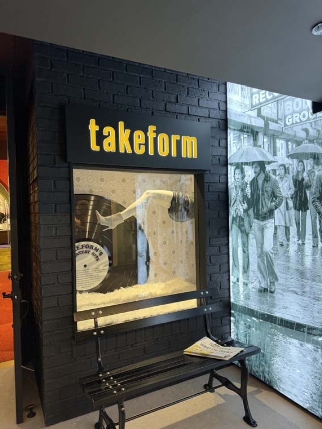 Black brick storefront labeled 'takeform' with a mannequin leg and vinyl record display behind glass, bench and vintage newspaper nearby.