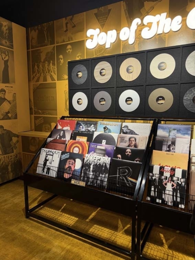 Vinyl display in warm yellow lighting: black metal racks holding albums beneath framed gold records and neon 'Top of The' sign.