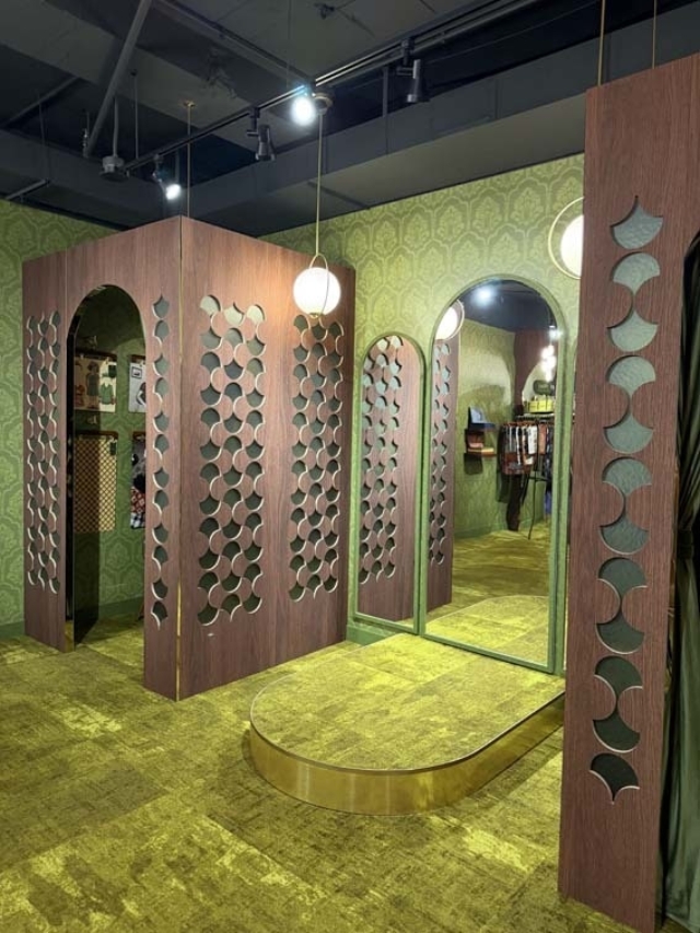 Retro fitting room with green wallpaper, gold carpet, arched mirror, wooden panels with circular cutouts and globe lights and racks.