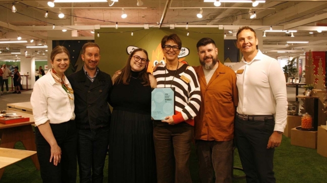Six smiling people stand shoulder-to-shoulder in a lit gallery under string lights, center person holding a blue 'WINNER' award plaque.