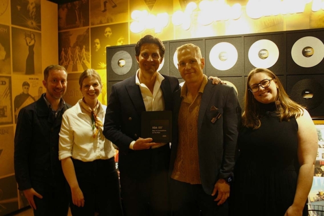 Five smiling adults stand in a warmly lit room with retro records on the wall, the center man holding a small award plaque flanked by peers.