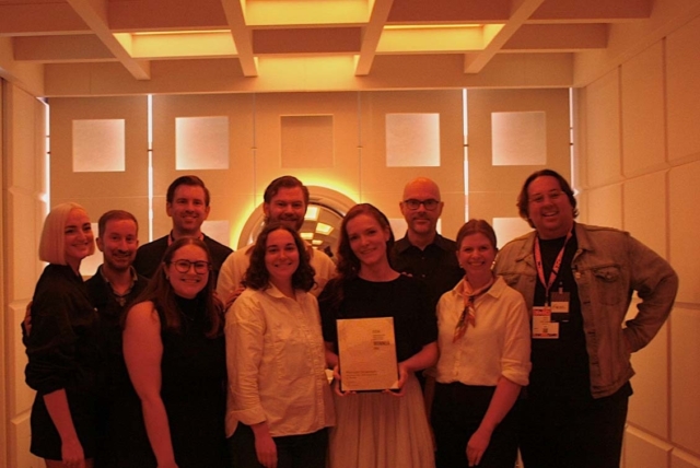 Eleven smiling adults stand in a warm amber-lit geometric room; a woman at center holds a framed award while colleagues cluster around her.