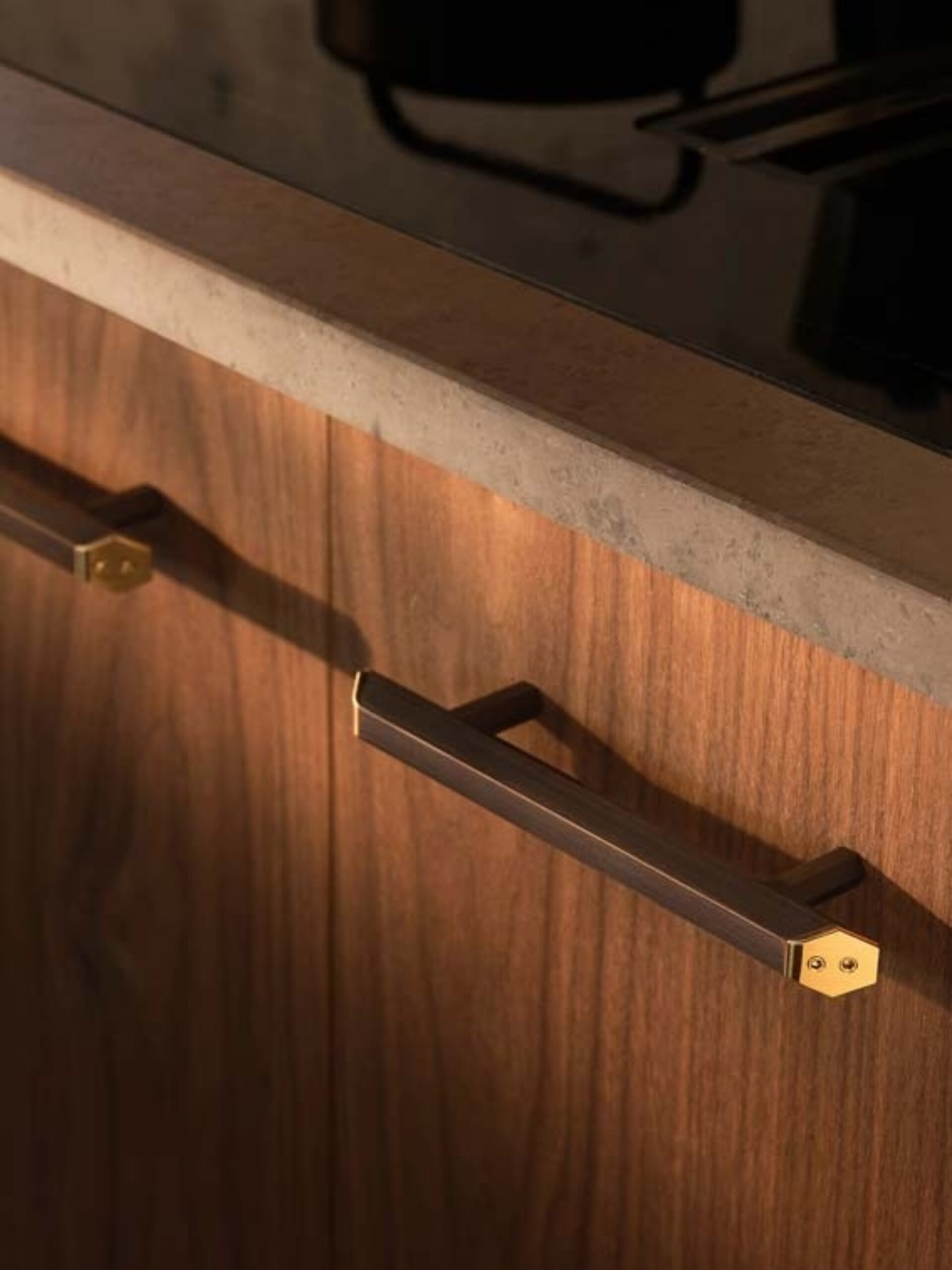 Warm walnut cabinet door under a stone countertop with a long dark bronze handle featuring hexagonal brass end caps and soft shadow.