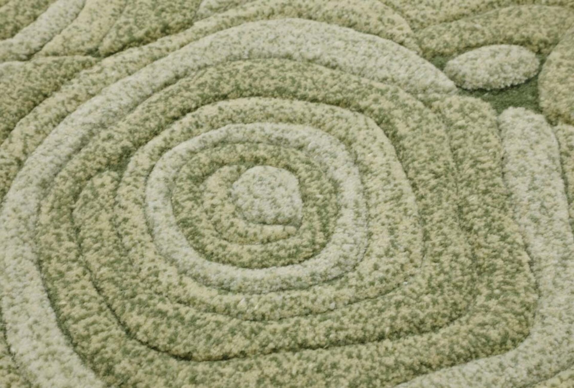 Closeup of a soft looped green and cream carpet with raised concentric rounded-square and spiral ridges creating a textured, organic pattern.