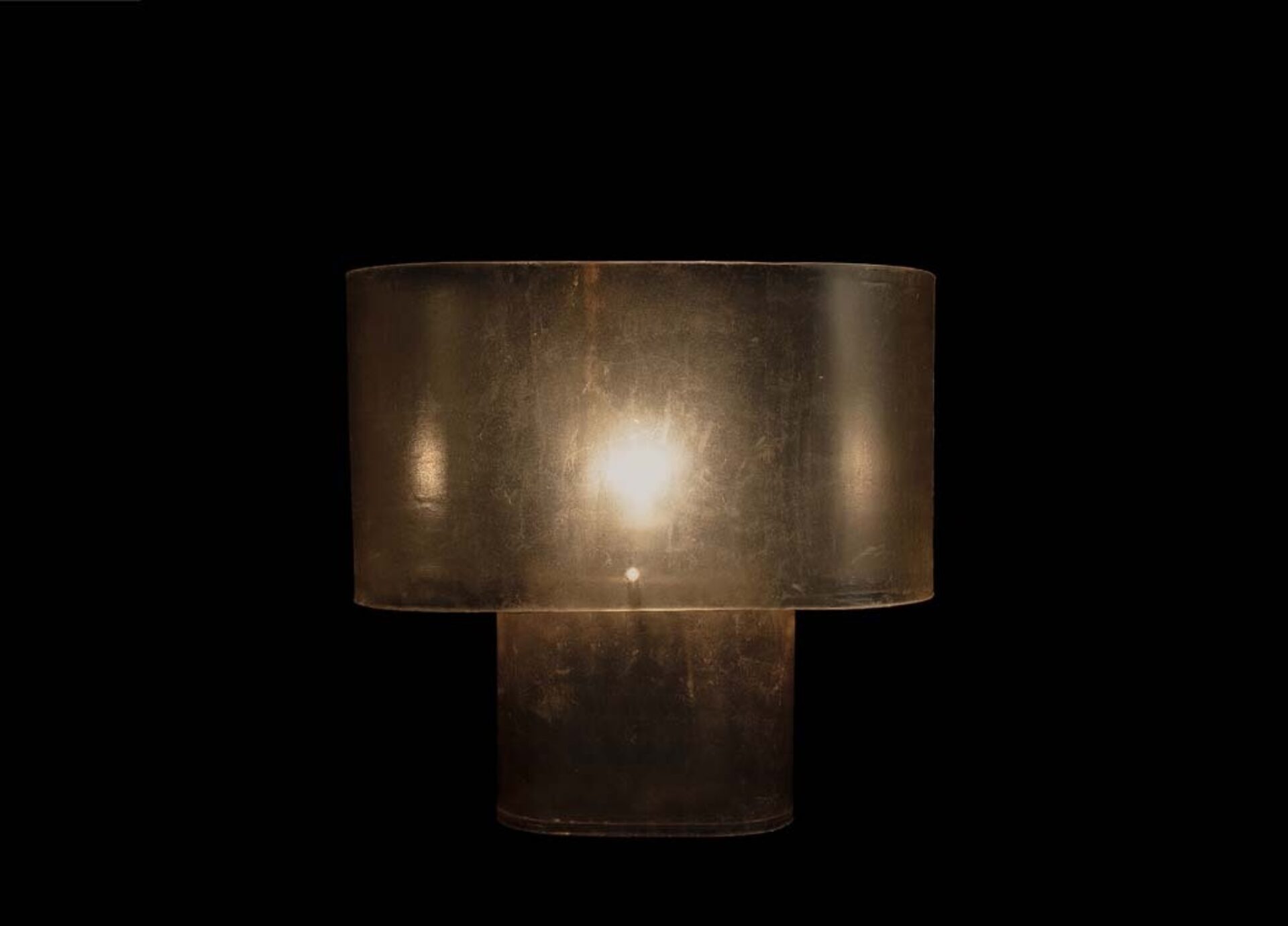 A warm glowing lamp with two translucent cylindrical sections, lightly scratched and dusty, casting soft amber light against black.