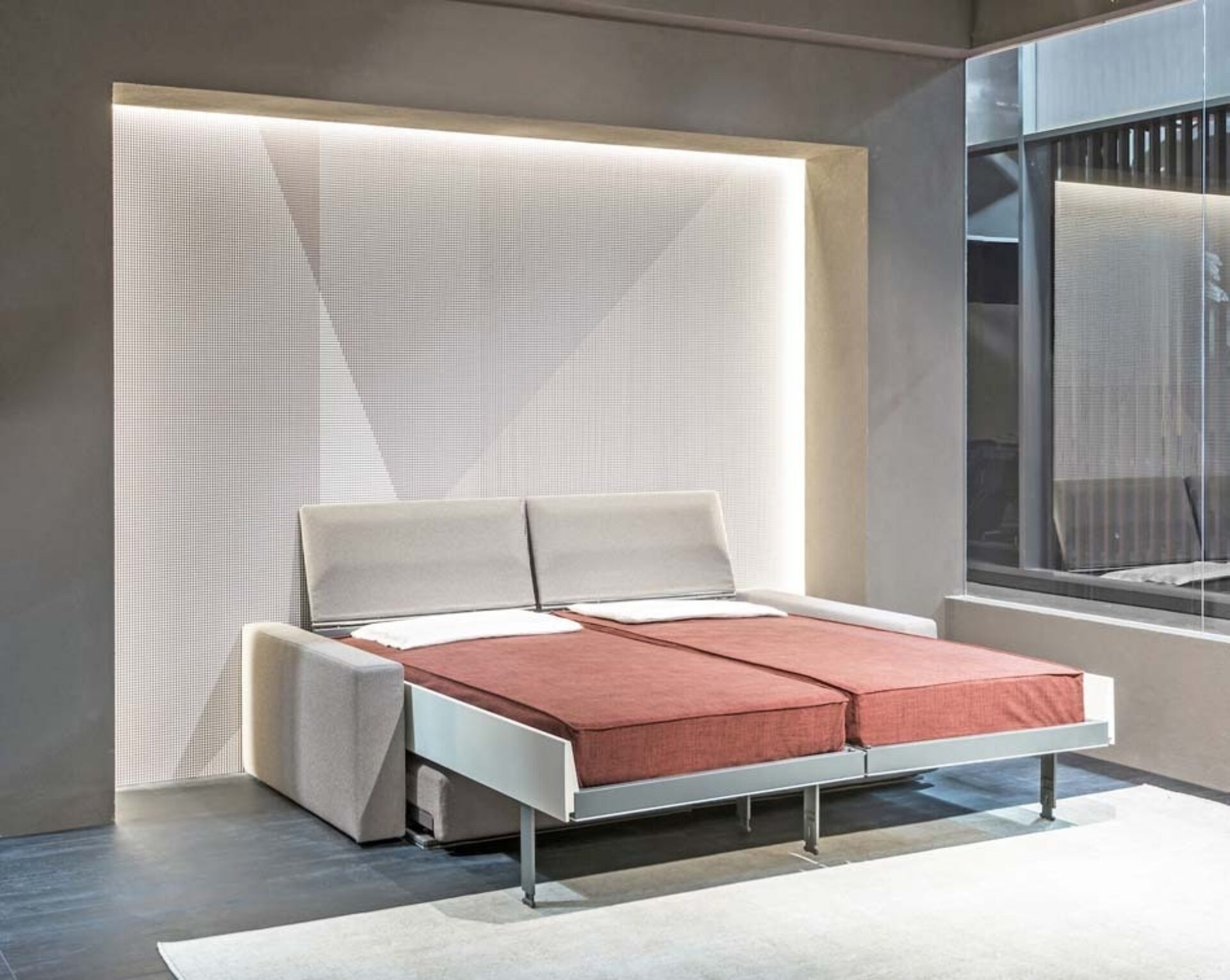 Light-gray sofa bed opened into two red twin mattresses against a backlit textured wall, beside a tall window and concrete floor.