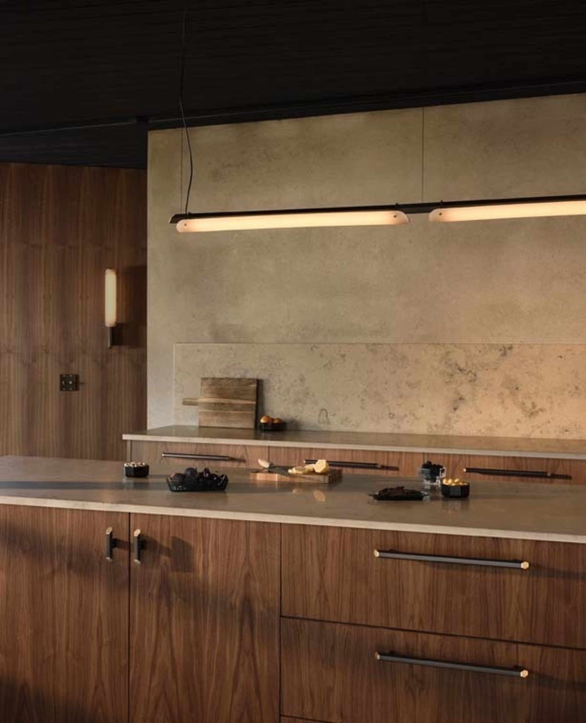 Warm modern kitchen with walnut cabinets, concrete backsplash, a long suspended tubular pendant light, marble countertop and small bowls.