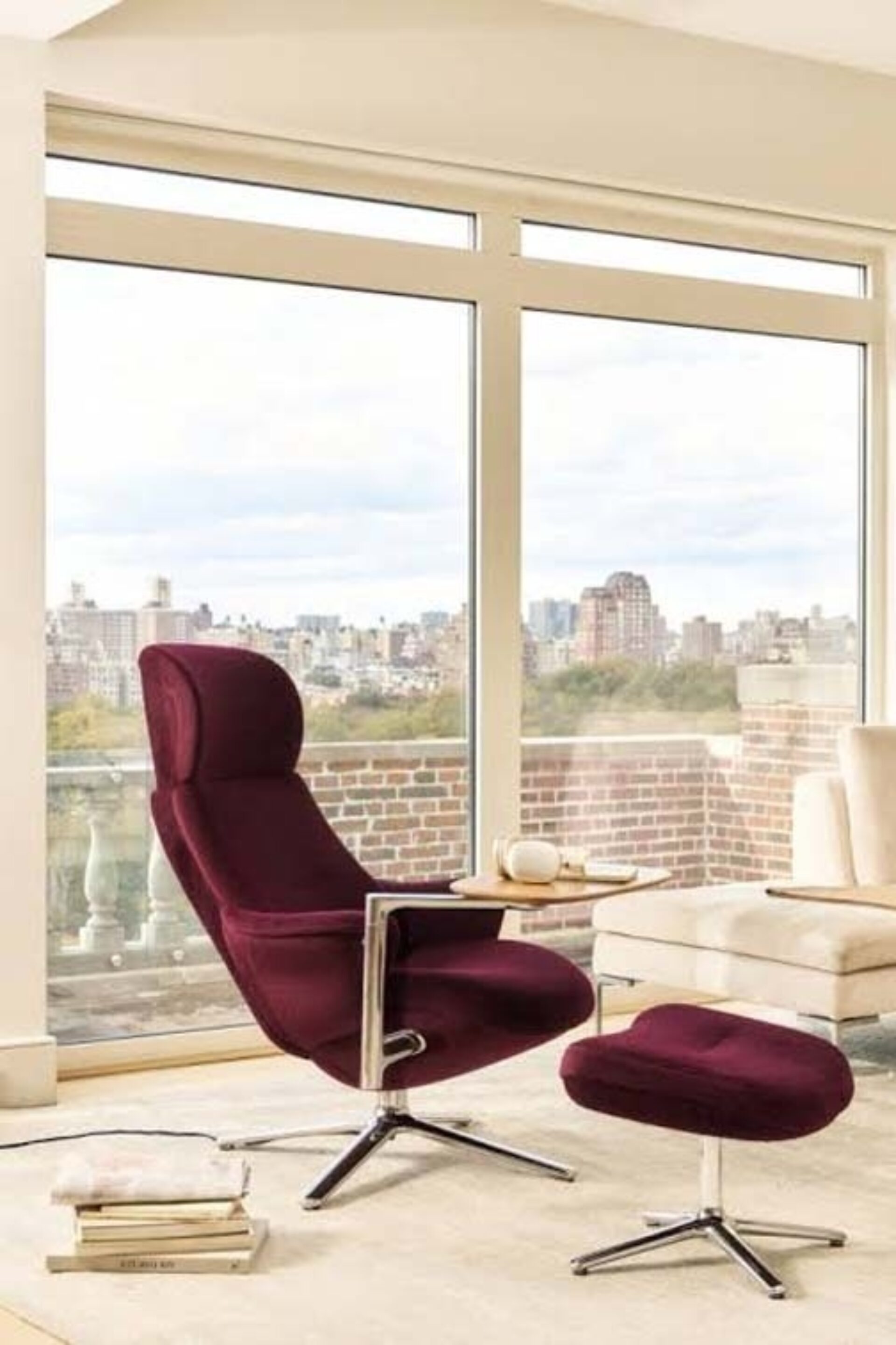 Burgundy swivel lounge chair with matching ottoman and chrome bases beside a side table and cream sofa in a sunlit city-view living room.