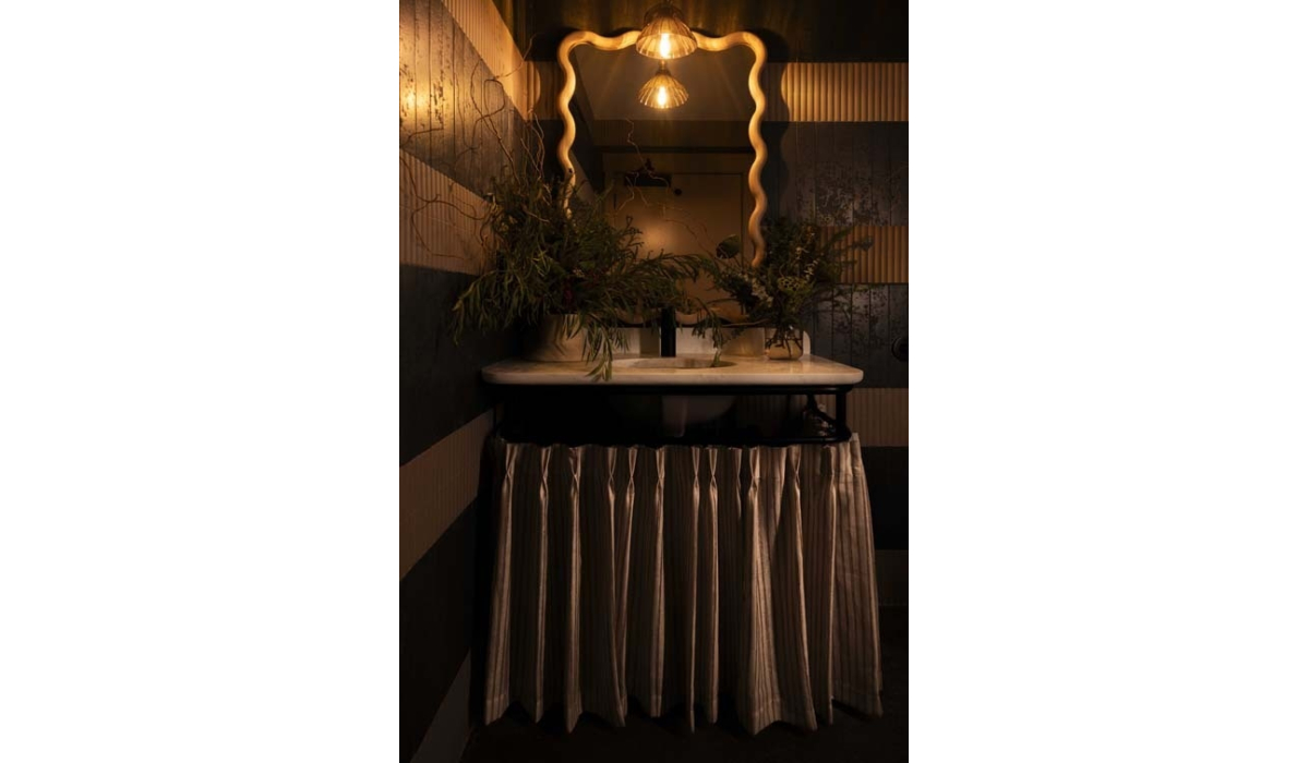 A stylish bathroom features a vintage sink with a curtain, ornate mirror, and warm lighting, complemented by floral arrangements.