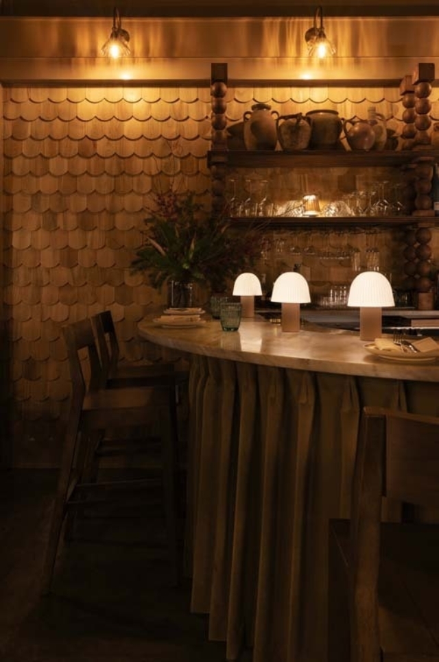 Soft, warm lighting casts a cozy glow over a bar with elegant decor, featuring round lamps, wooden shelves, and inviting seating.