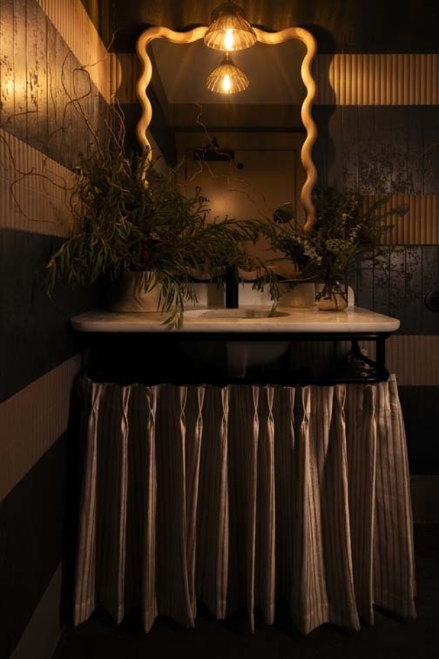 A stylish bathroom features a vintage sink with a curtain, ornate mirror, and warm lighting, complemented by floral arrangements.