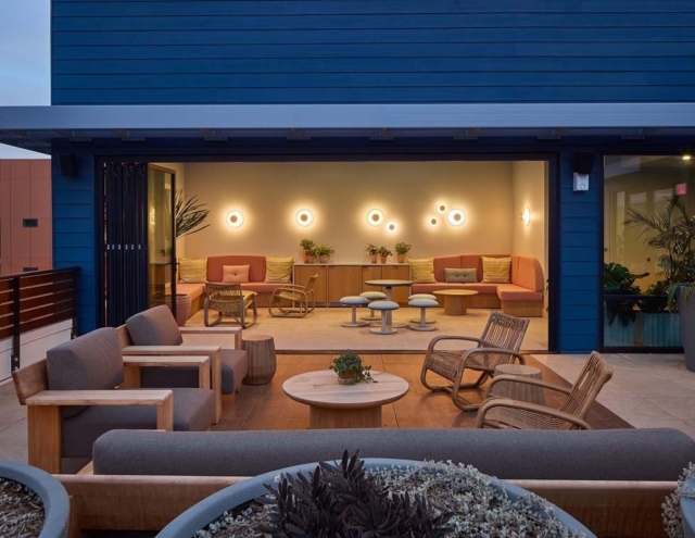 A modern outdoor lounge features wooden furniture, soft seating, and stylish lighting, surrounded by potted plants for a cozy atmosphere.