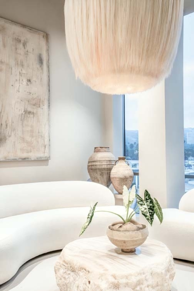 A modern living space features a plush white sectional, a round natural wood coffee table, ceramic vases, and a large pendant light above.
