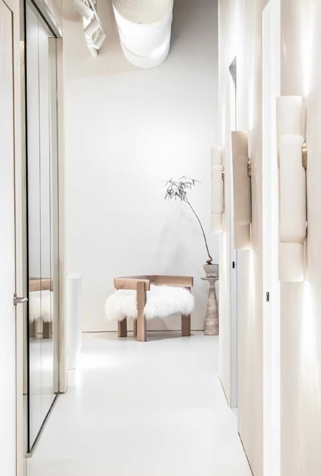 A minimalist hallway features a mirrored closet door, soft lighting, a modern wooden bench with a fur seat, and a decorative plant.