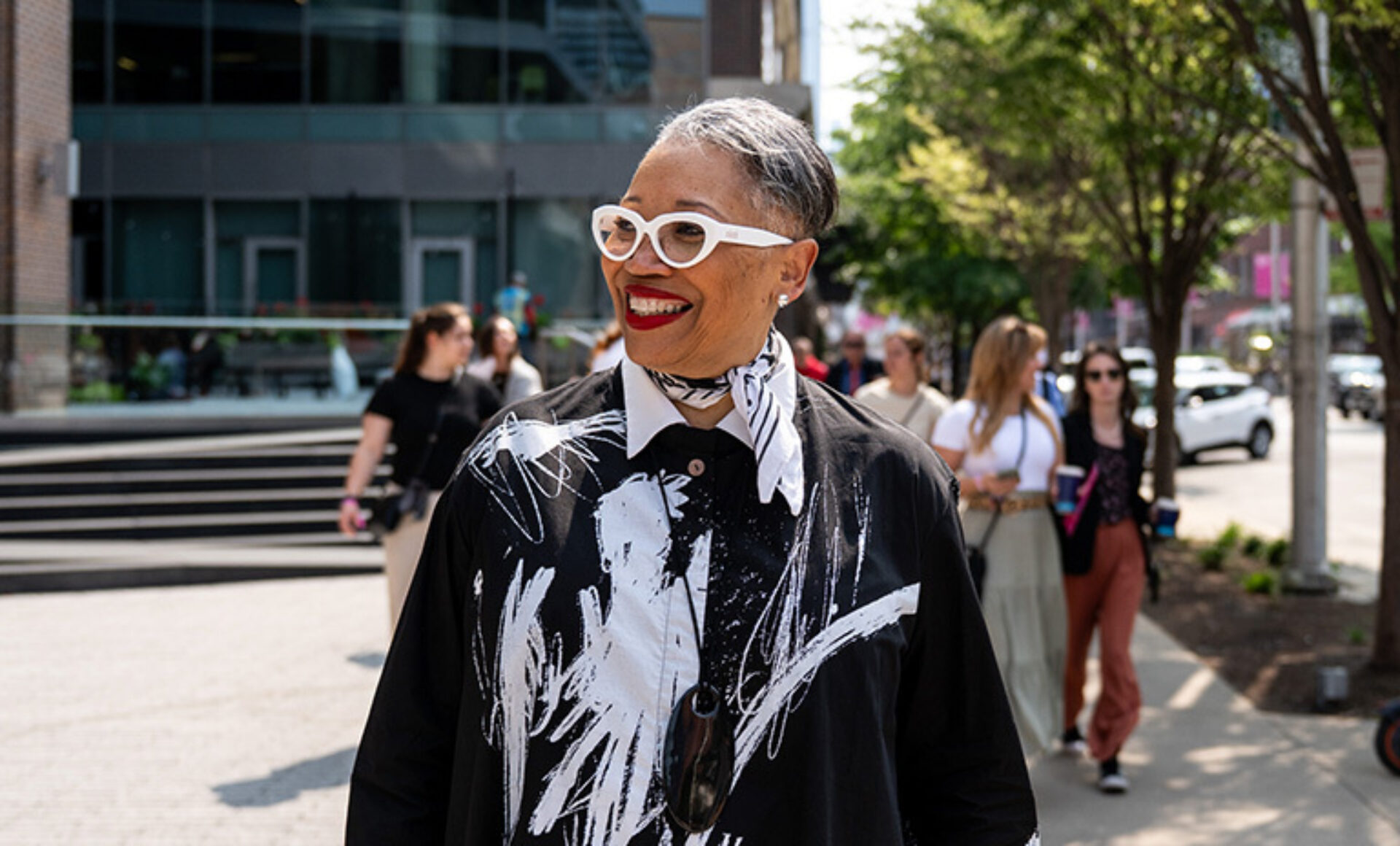 A woman with stylish white glasses and a vibrant, graphic black-and-white outfit smiles while walking in a bustling city street.