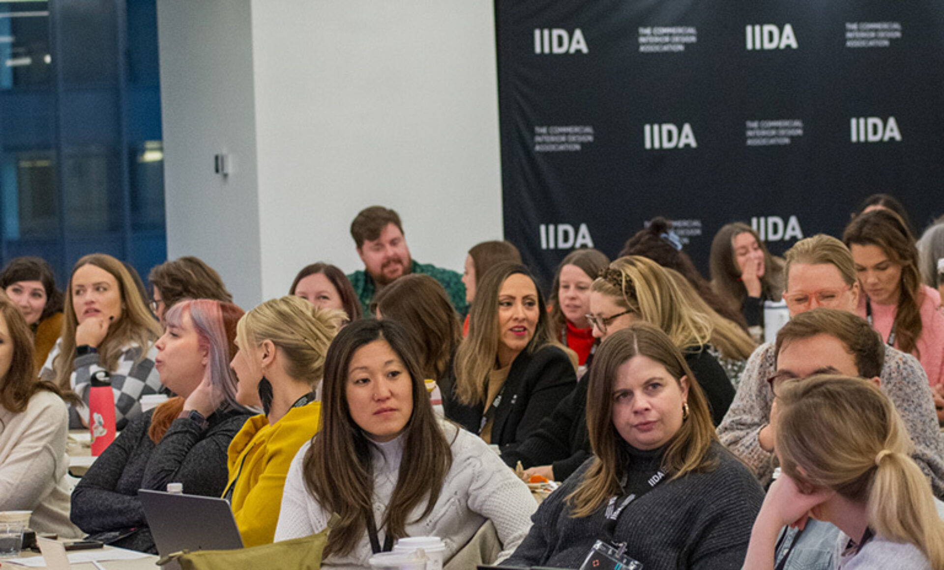 A diverse group of people, mostly women, engaged in conversation during an event, with a professional backdrop featuring the IIDA logo.
