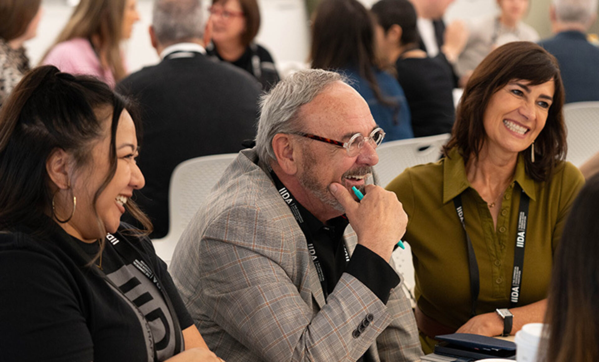 Three colleagues seated at a conference table laugh together; a gray-haired man in a checked blazer holds a pen while two women smile.