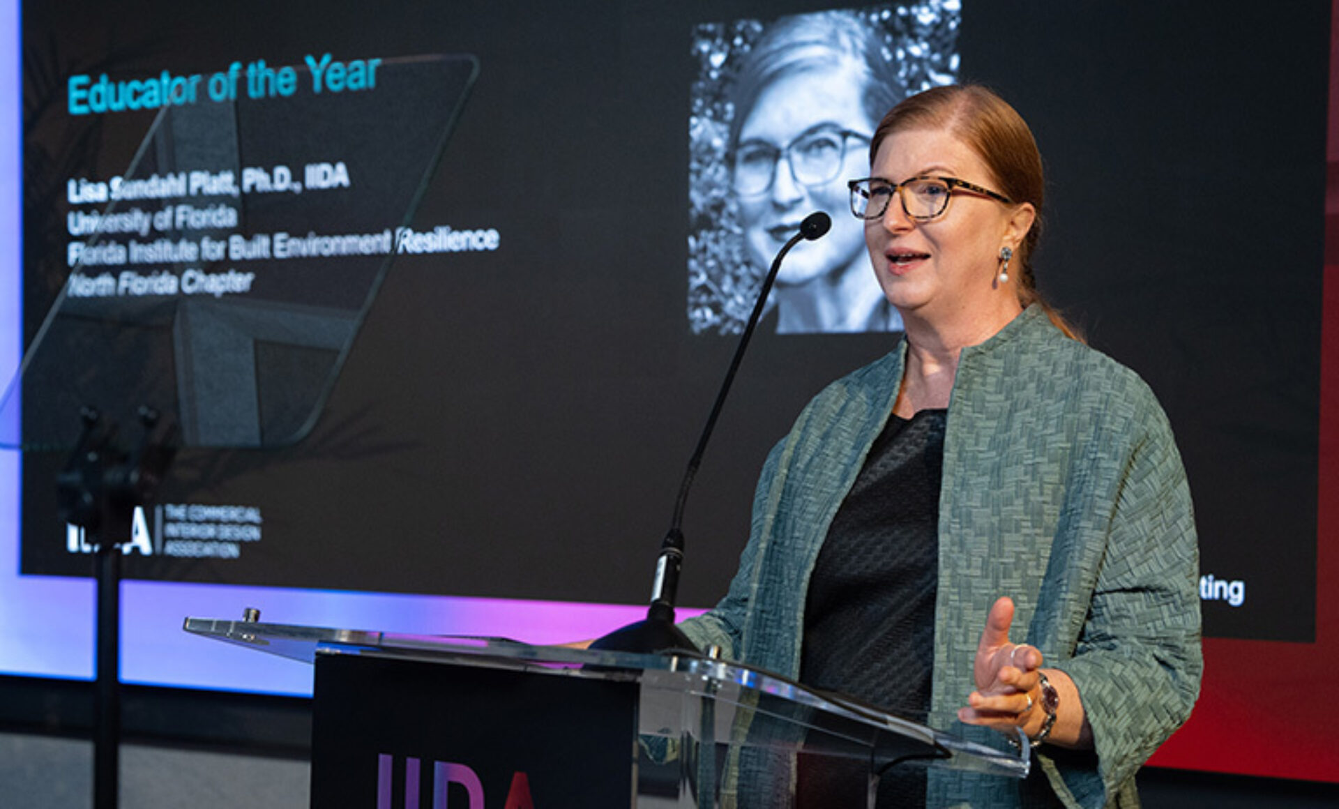 Red-haired woman with glasses speaking at a clear lectern, gesturing while a slide titled 'Educator of the Year' displays behind her.