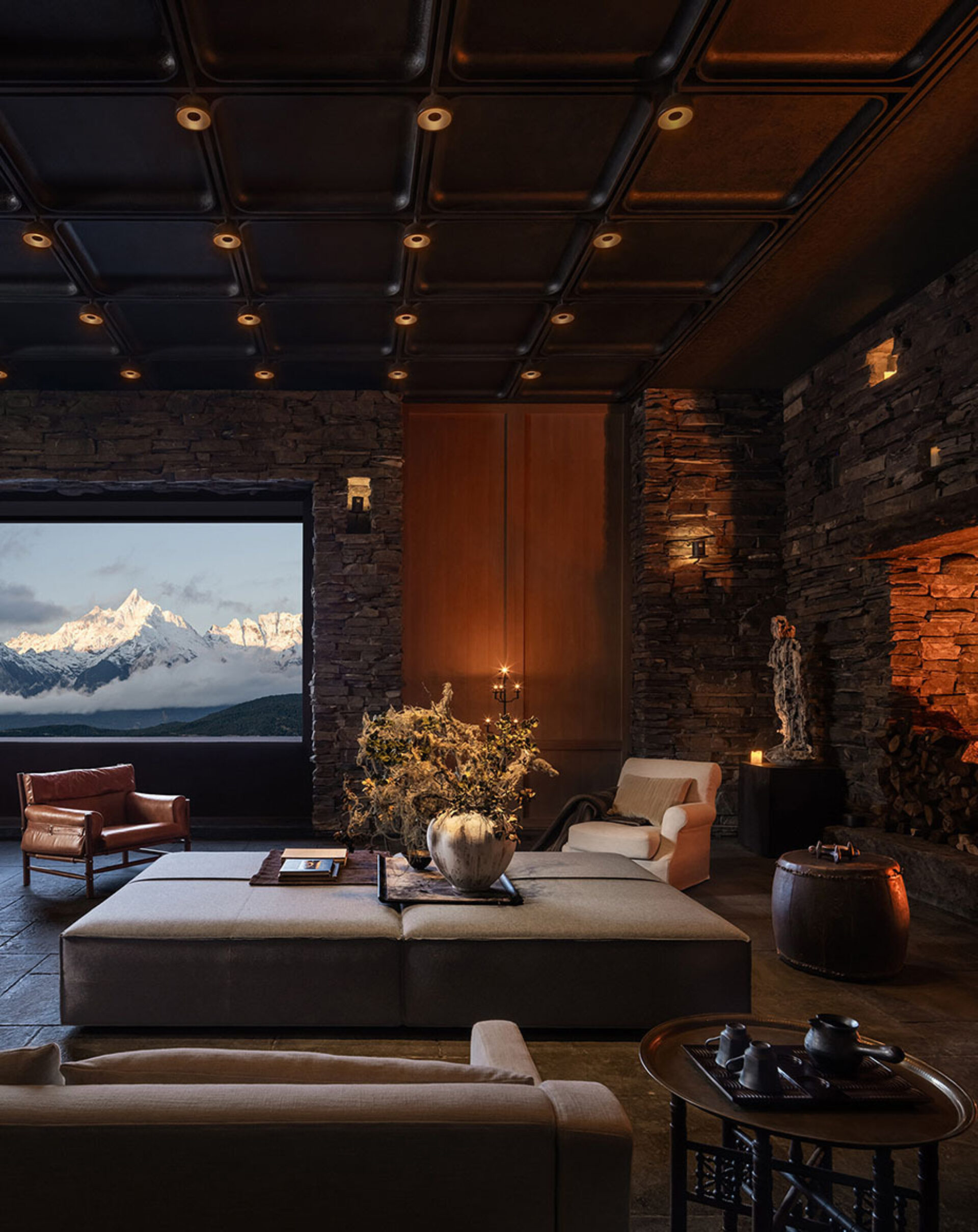 Cozy dimly lit stone-walled living room with low grey sectional, leather chair, vase of dried branches and window framing snow‑capped peaks.