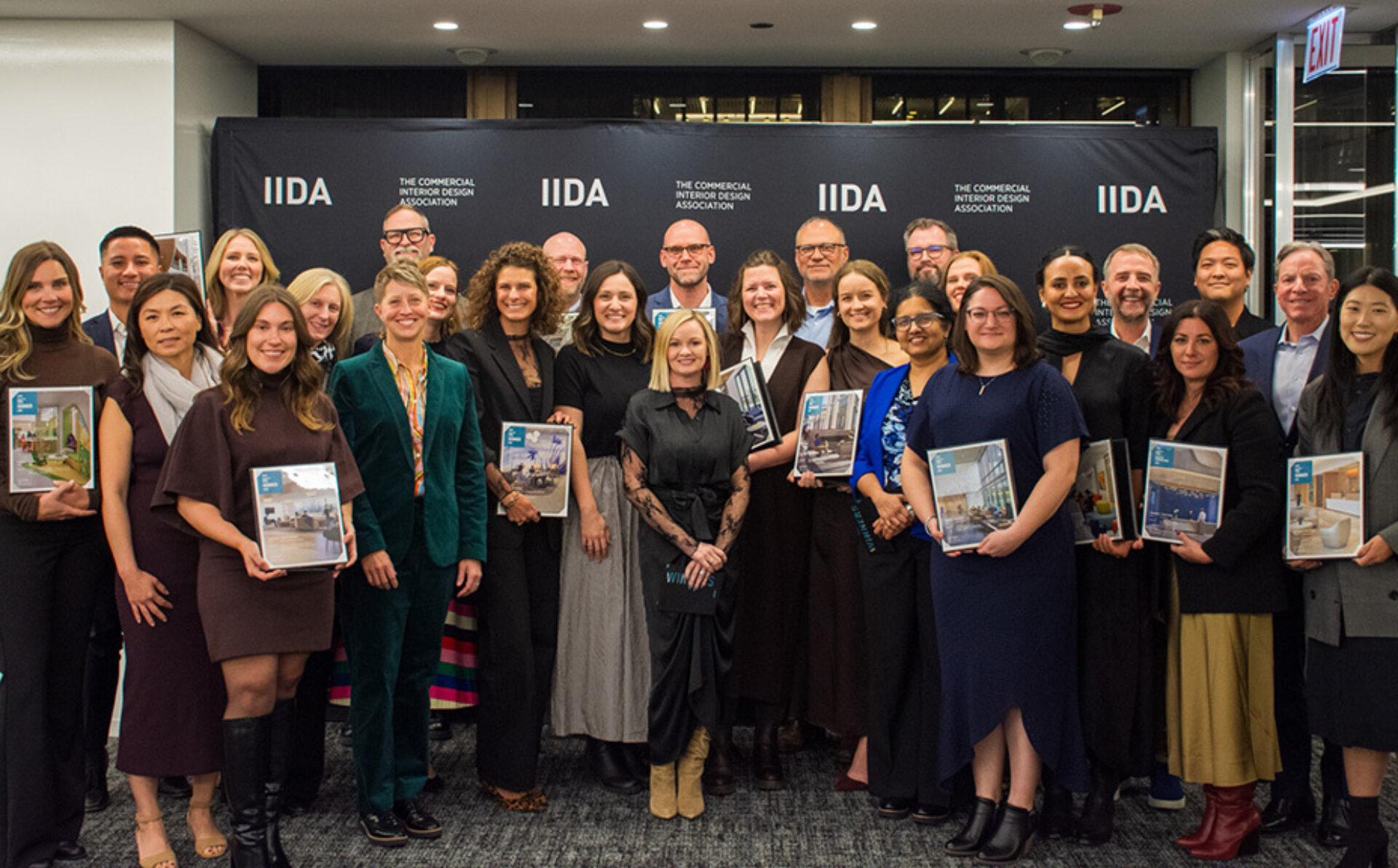Large diverse group of smiling interior designers and award winners posed in front of an IIDA backdrop, holding framed certificates.