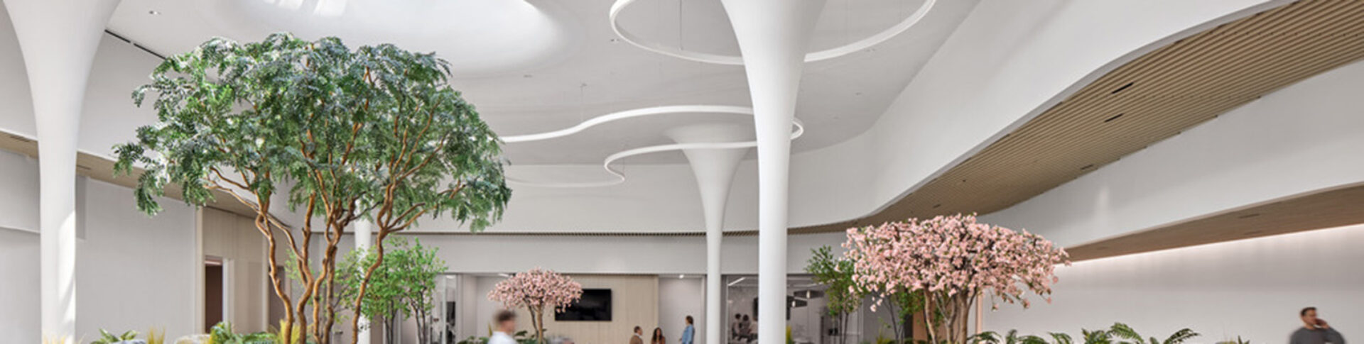 Bright modern atrium with white funnel columns, curved ceiling lights, tall green and pink blossoming trees, planters and blurred people.