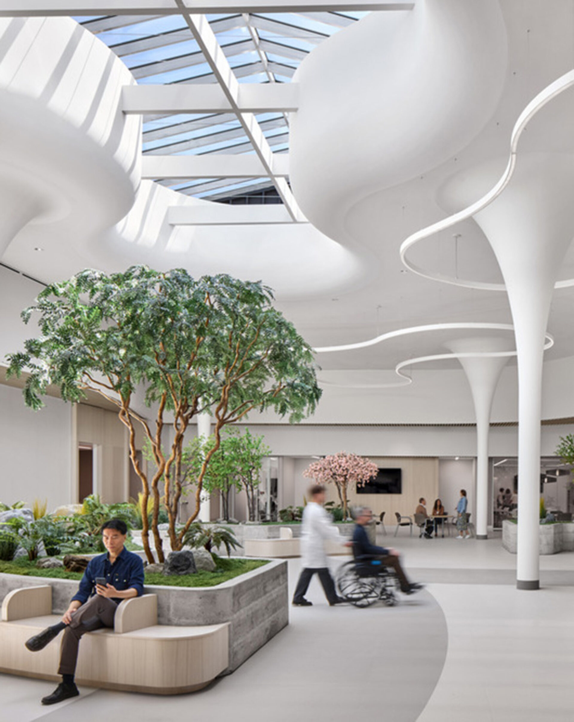Bright modern hospital atrium with curving white columns, skylight, indoor trees and plants, a man seated, and a nurse pushing a wheelchair.