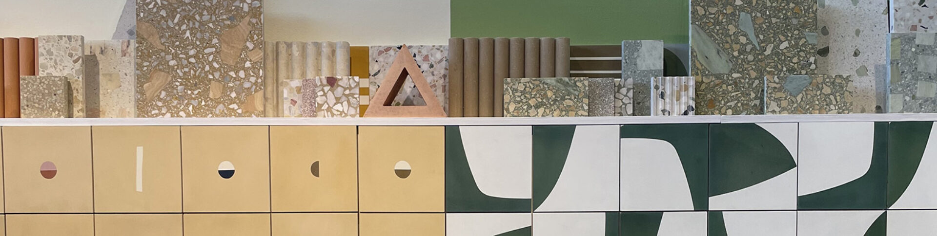 Shelf display of terrazzo and ceramic samples above mustard cubby fronts with circular cutouts and green-and-white abstract tile panels.
