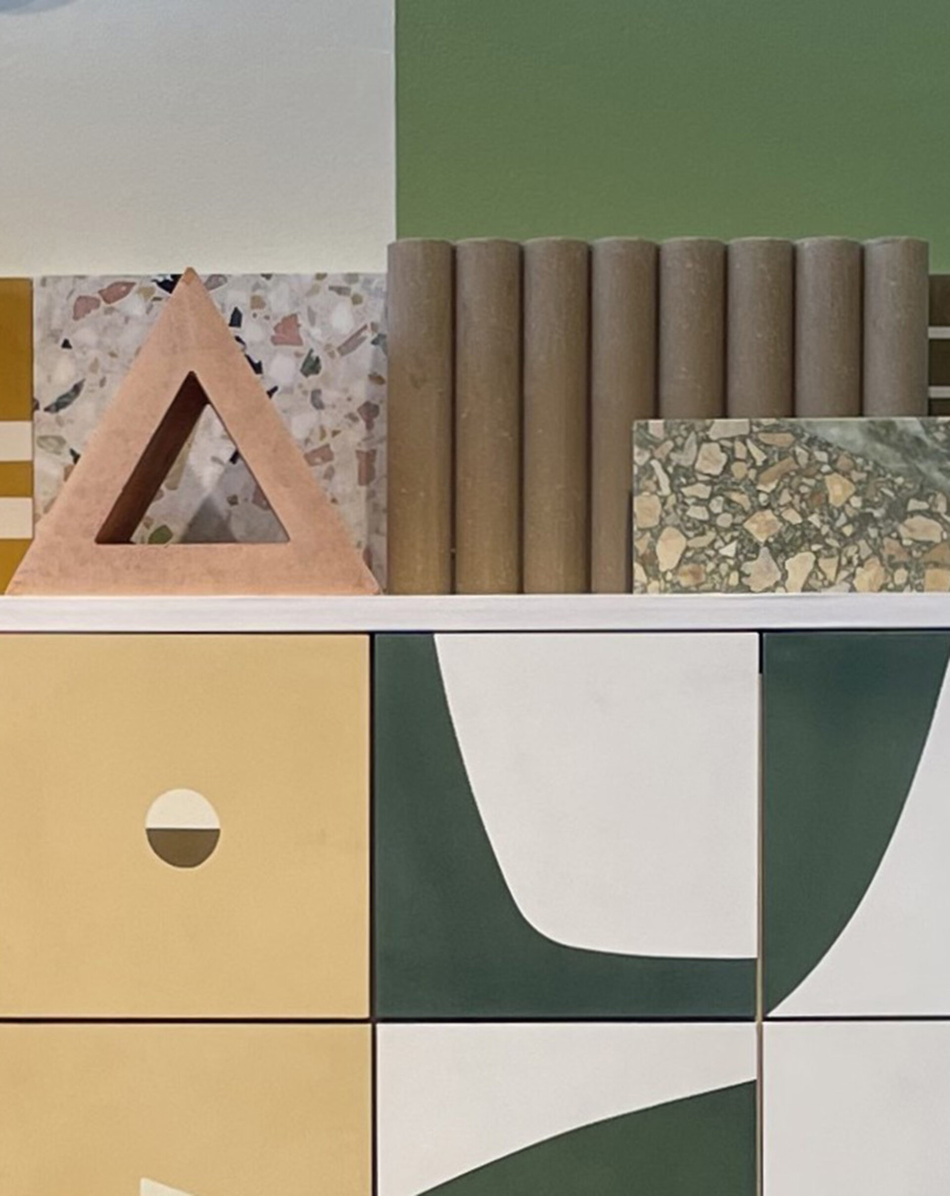Design samples on a geometric cabinet: mustard, white and green panels, terrazzo slabs, hollow terracotta triangle and brown tubes.