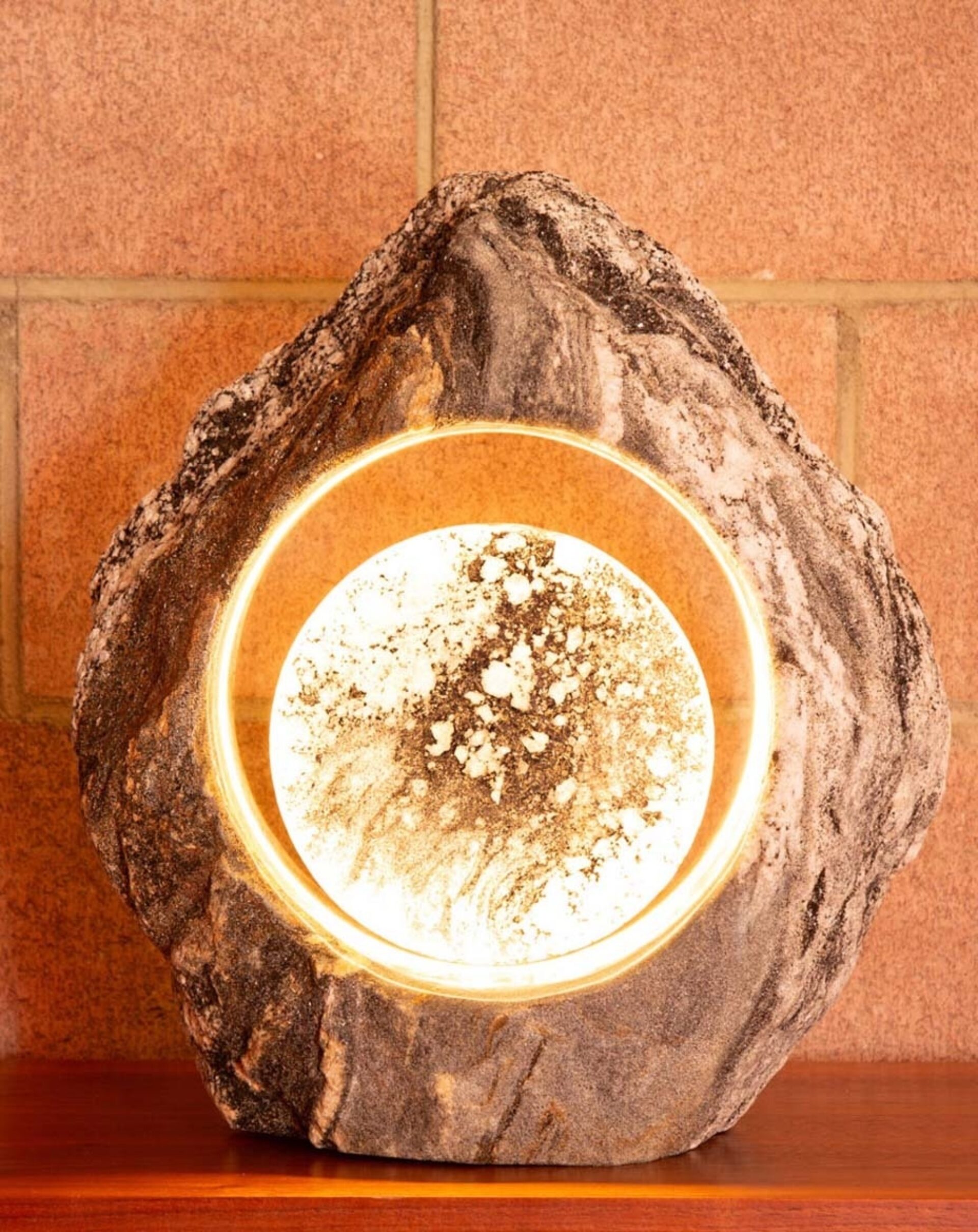 Warm amber orb recessed in a rough marbled gray stone, softly glowing like a small sun against terracotta tiles on a wooden shelf.
