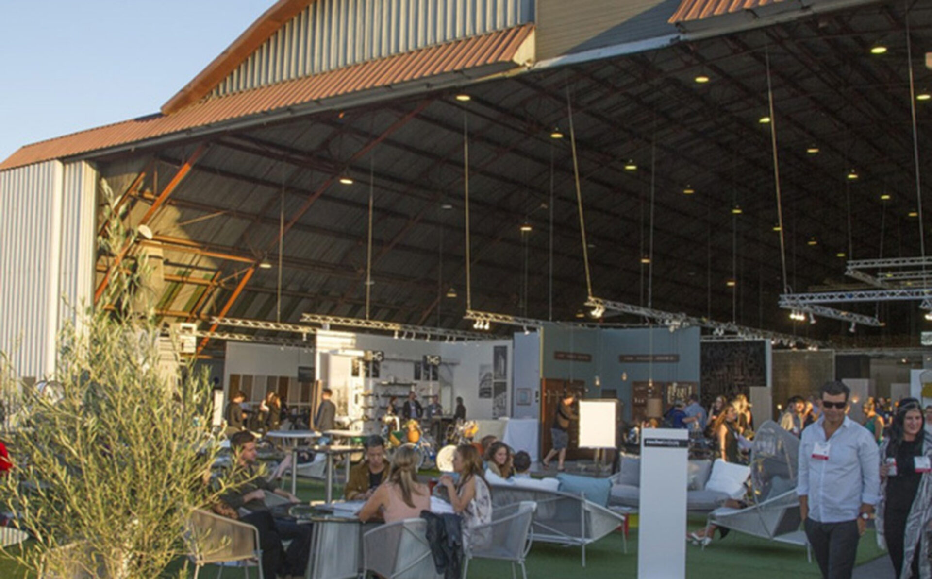 Spacious hangar converted into a lounge event with groups chatting on sofas, tables, greenery and overhead lighting rigs, at dusk.
