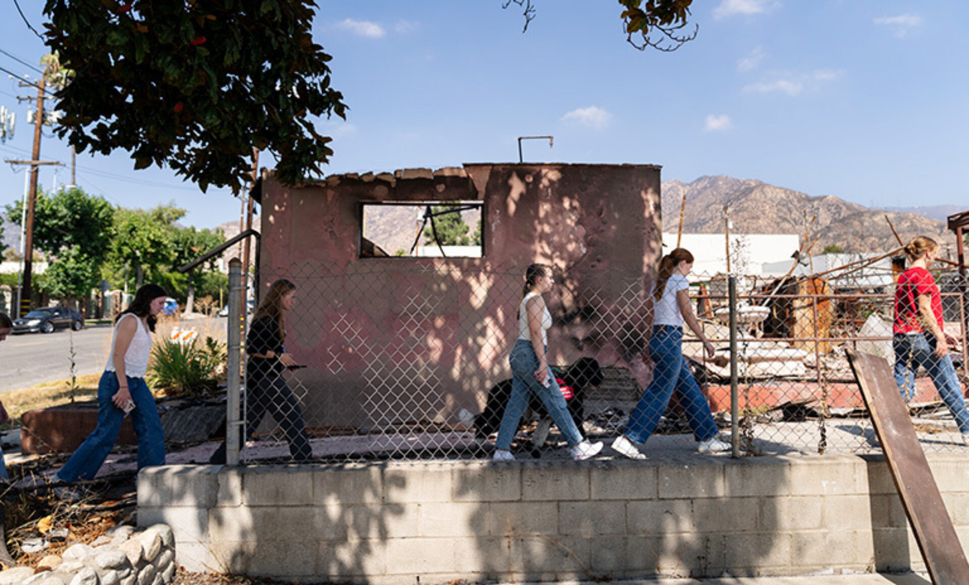 Five casually dressed pedestrians walk past a chain-link fence in front of a scorched, windowless pink structure with mountains behind them.