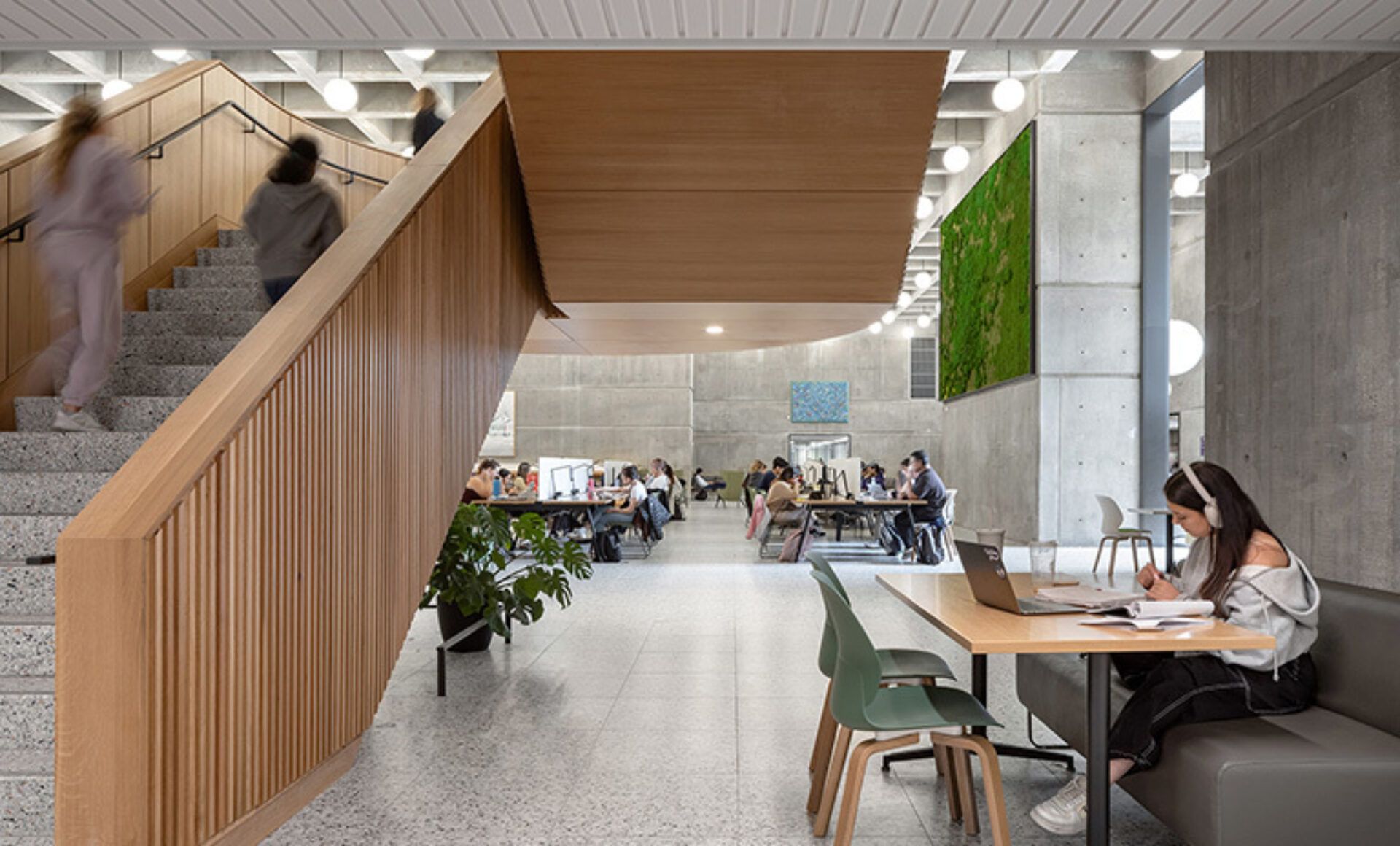Modern concrete-walled study hall with a wooden staircase, students at communal tables, moss wall art, and a woman with headphones studying.