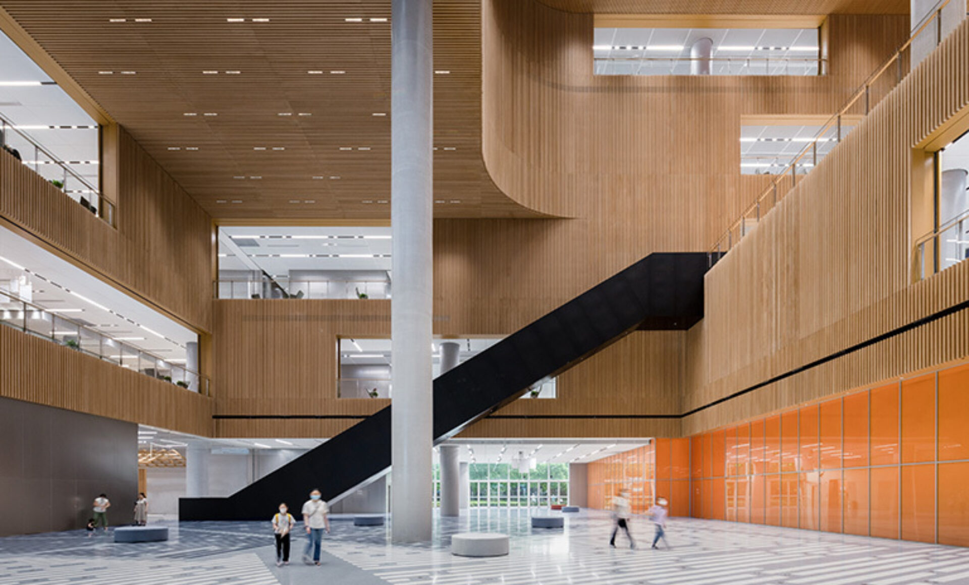 A spacious modern interior features large wooden walls, a striking black staircase, and people walking beneath bright overhead lights.