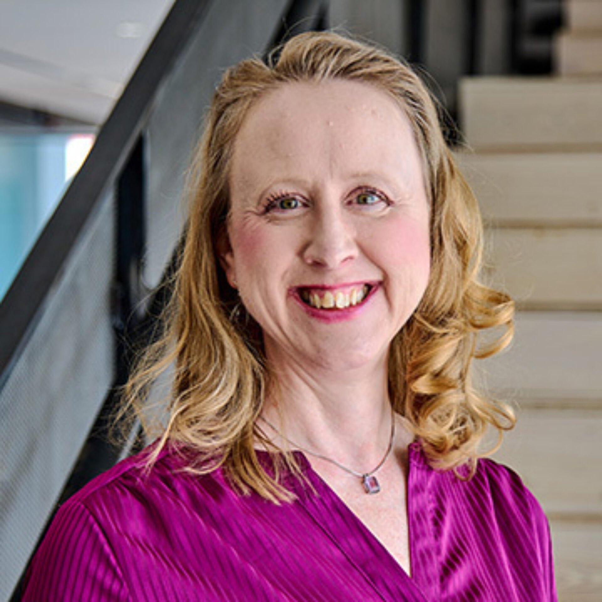 Smiling middle-aged woman with shoulder-length wavy blonde hair and magenta blouse, wearing a small pendant necklace, standing by stairs.