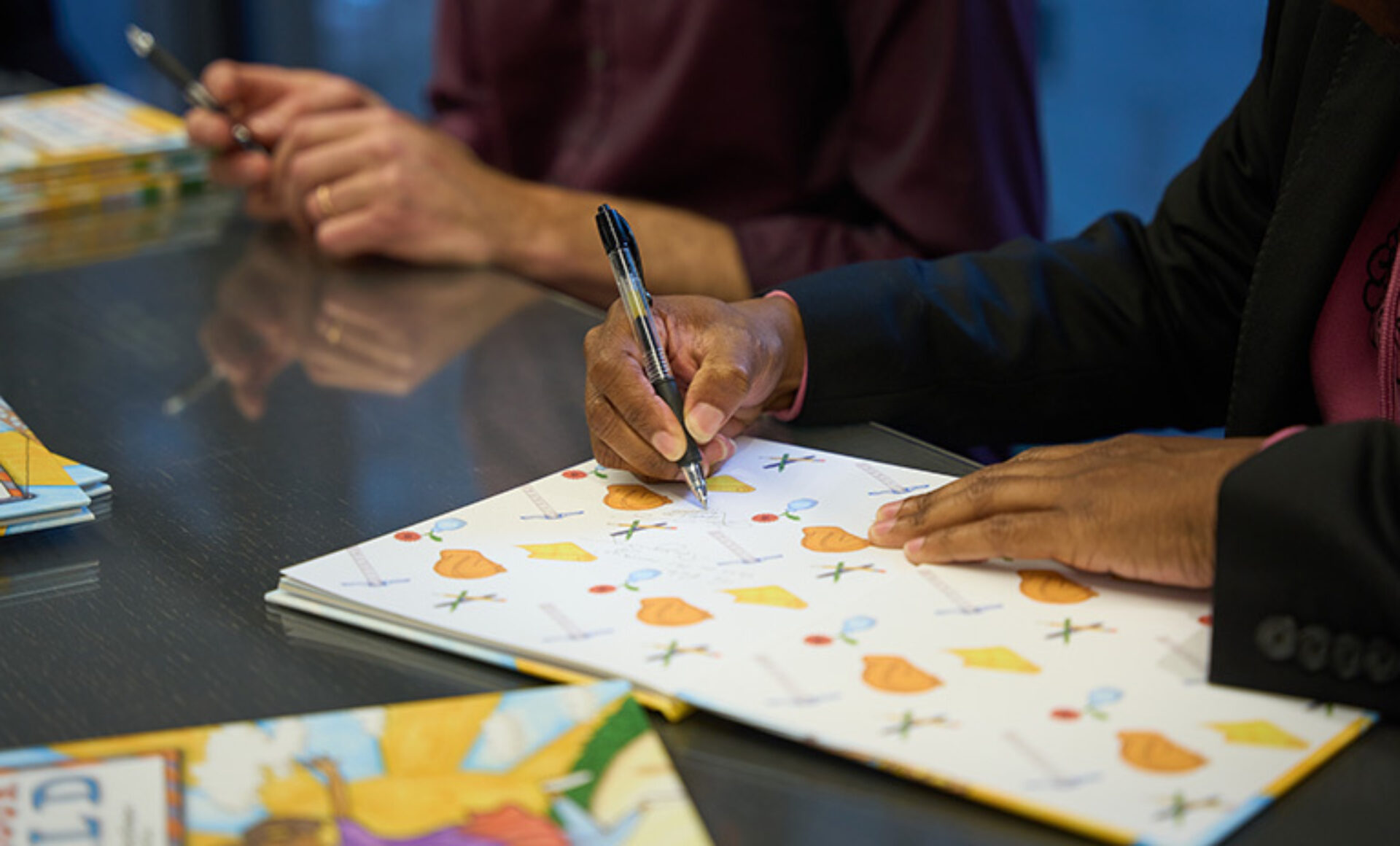 A hand holds a pen, signing a colorful page adorned with illustrations, while another hand rests on the table nearby.
