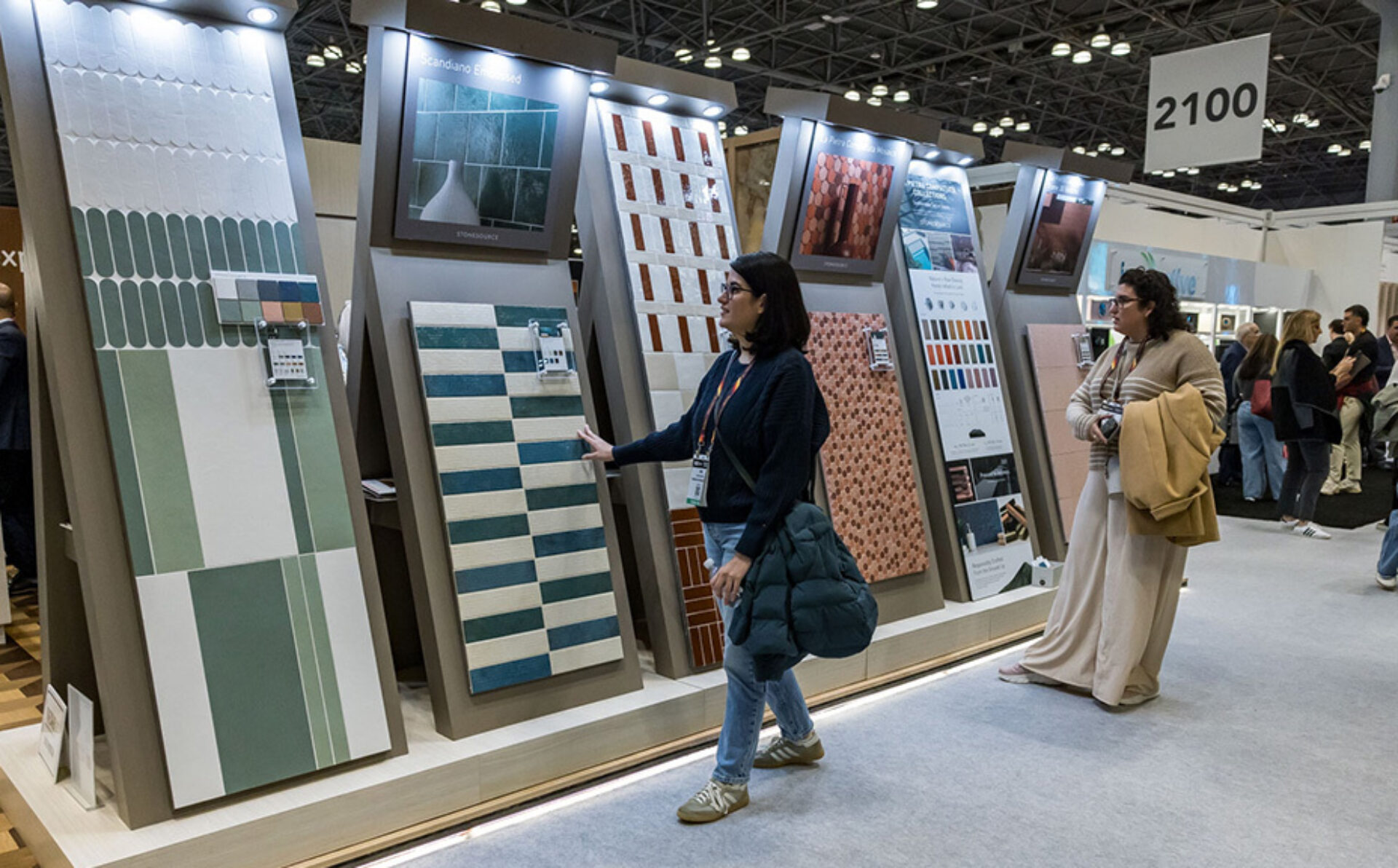 Two women examine large vertical tile sample boards at a busy trade show booth, one reaching to touch a green striped tile panel.