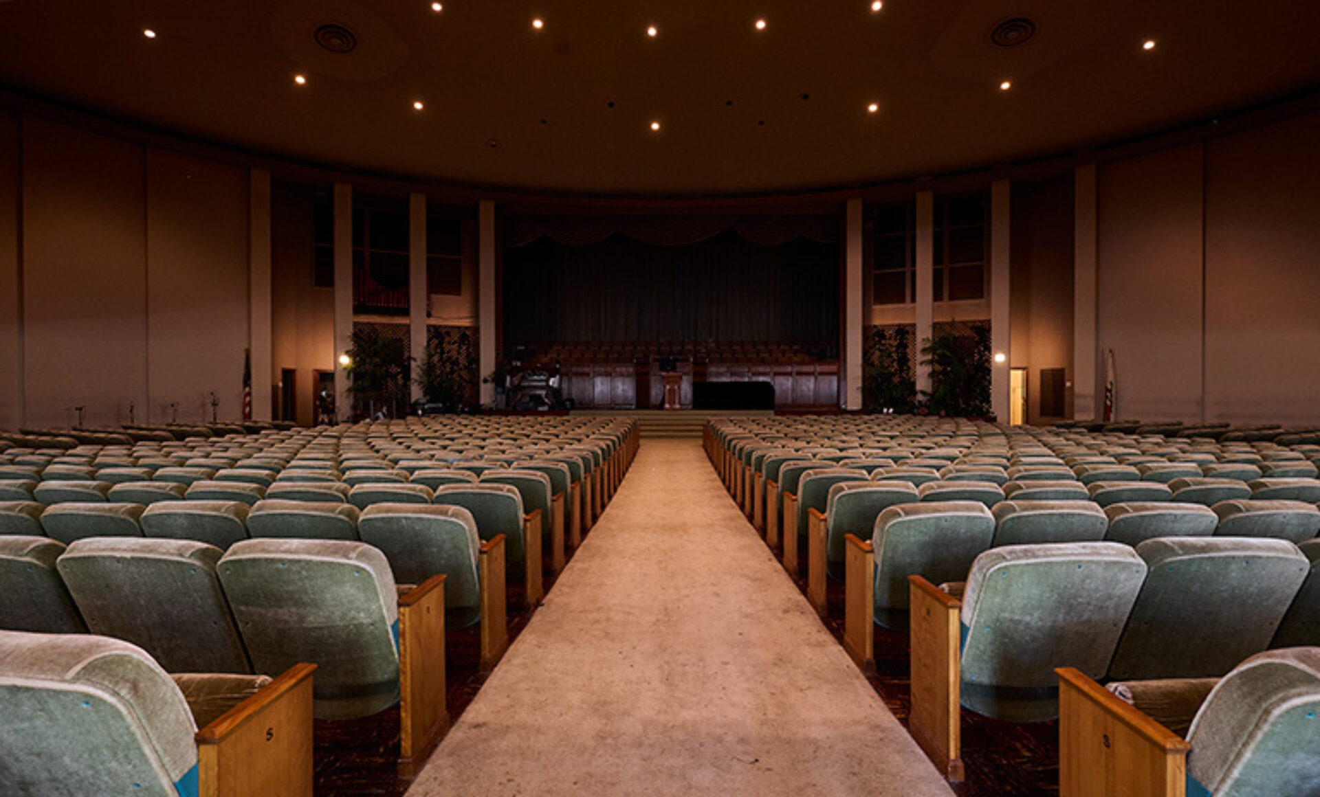 A spacious auditorium features rows of green seats, with a central aisle leading to a stage adorned with curtains and greenery, evoking nostalgia.