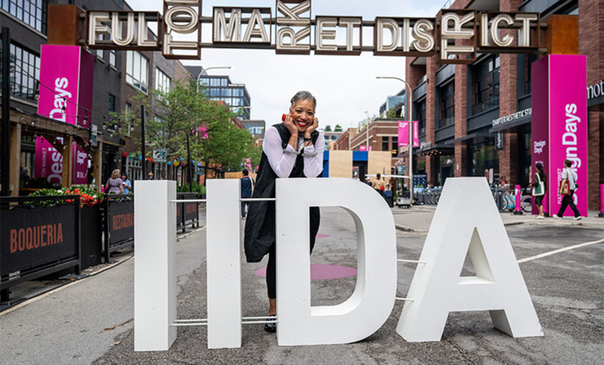 Smiling older woman with gray hair leans on large white letters spelling HIDA under a Fulton Market sign during the Design Days festival.