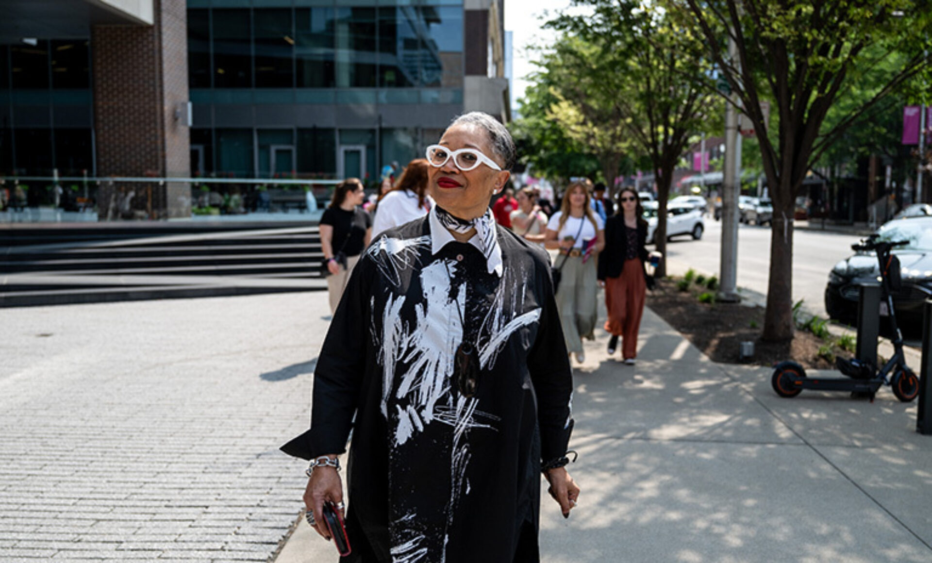 A stylish older woman with glasses and a bold black and white outfit walks confidently on a city street, surrounded by passersby.