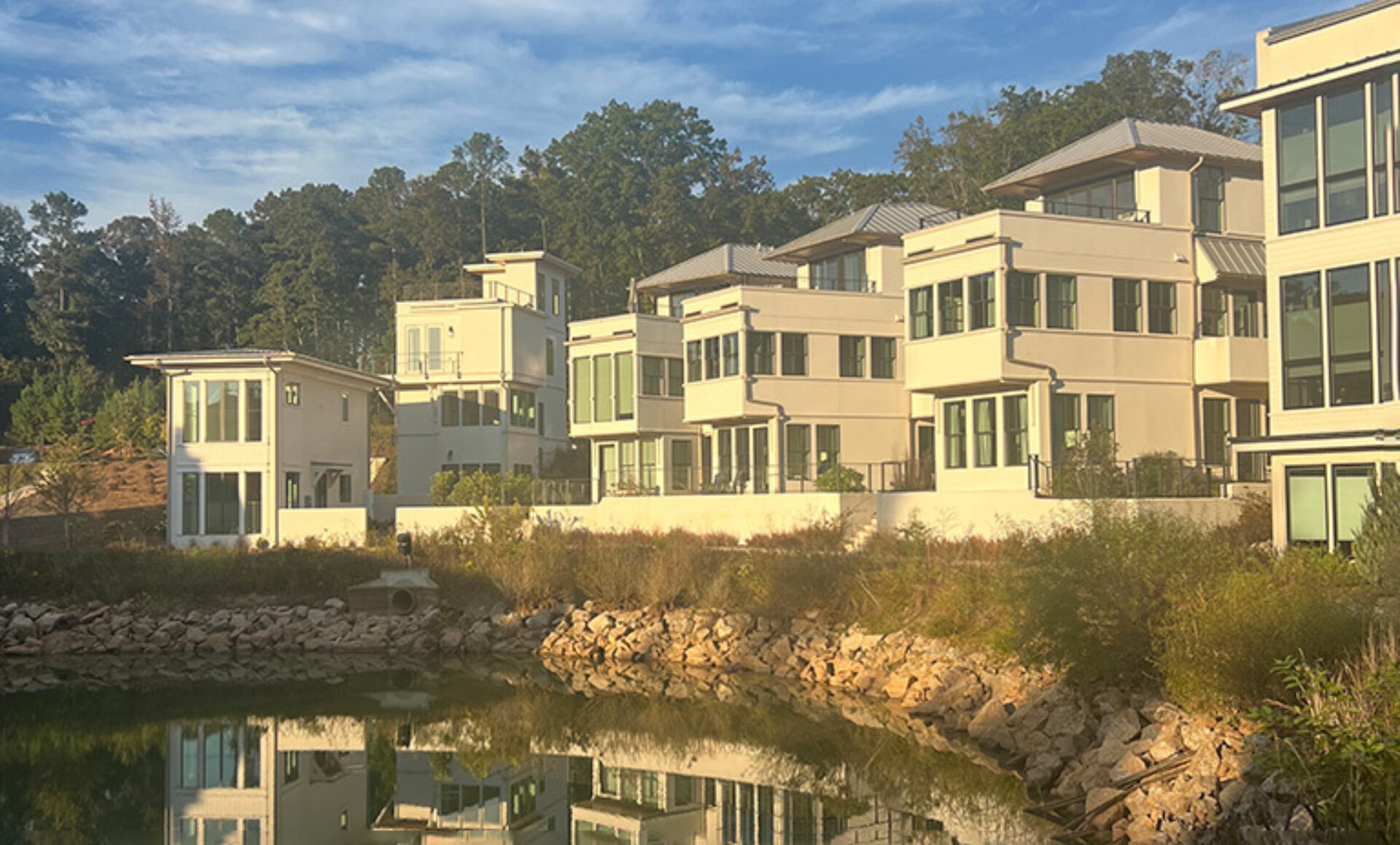Modern waterfront homes with multiple stories are reflected in a calm lake, surrounded by greenery and rocky shoreline.