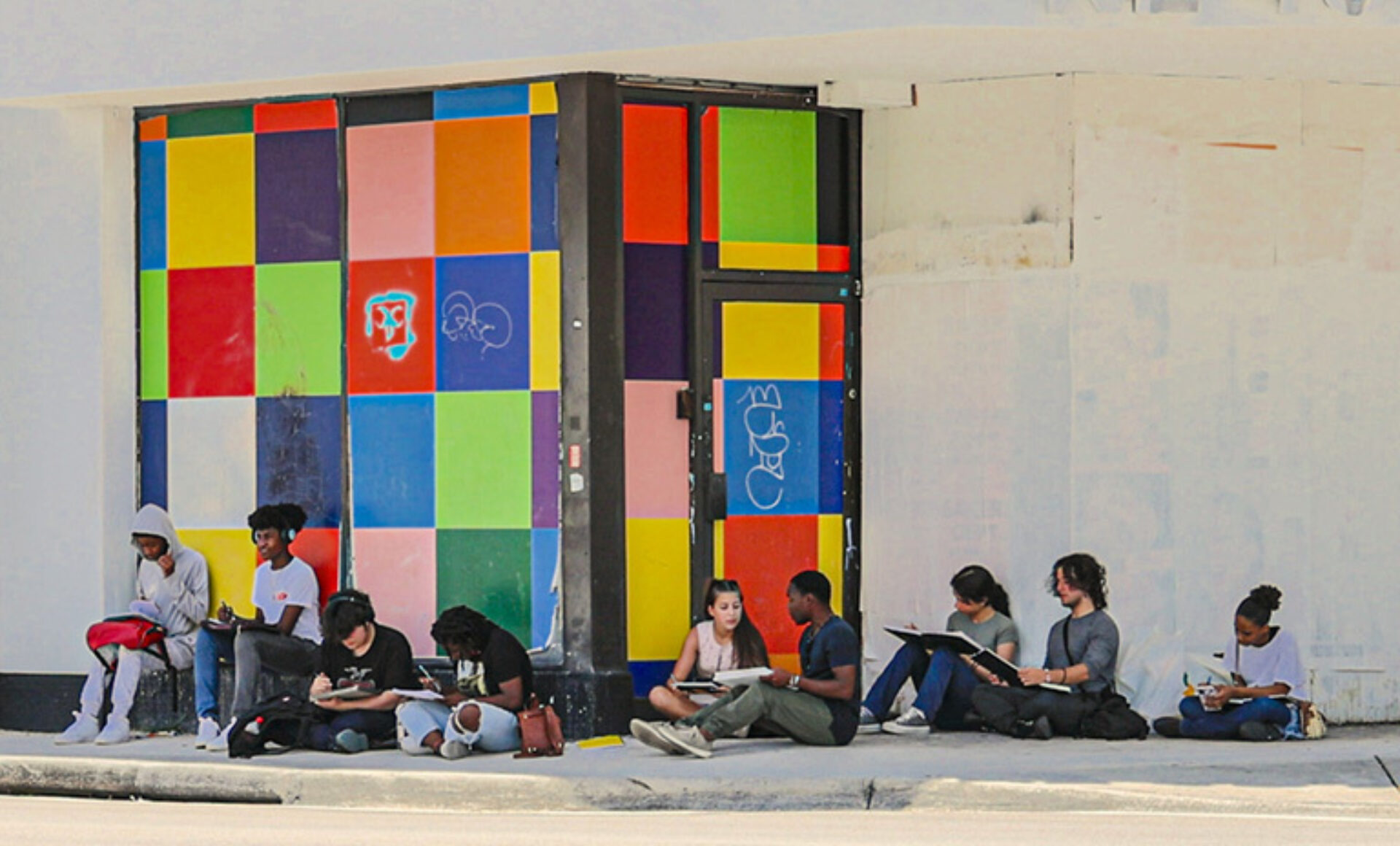 A diverse group of artists sits on the sidewalk, sketching in notebooks, next to a vibrant, colorful wall of squares.