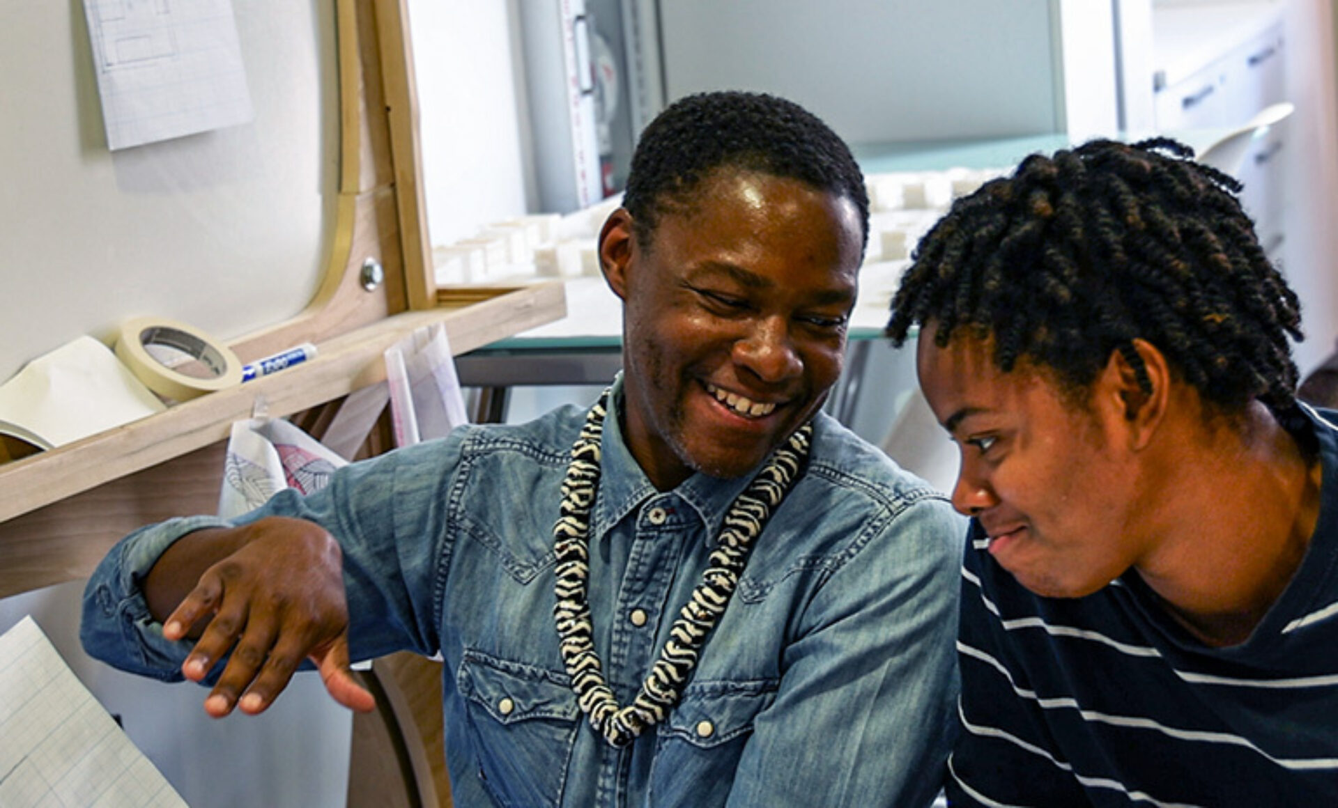 Two young men sit close together, smiling and engaged in conversation, surrounded by design materials in a creative workspace.