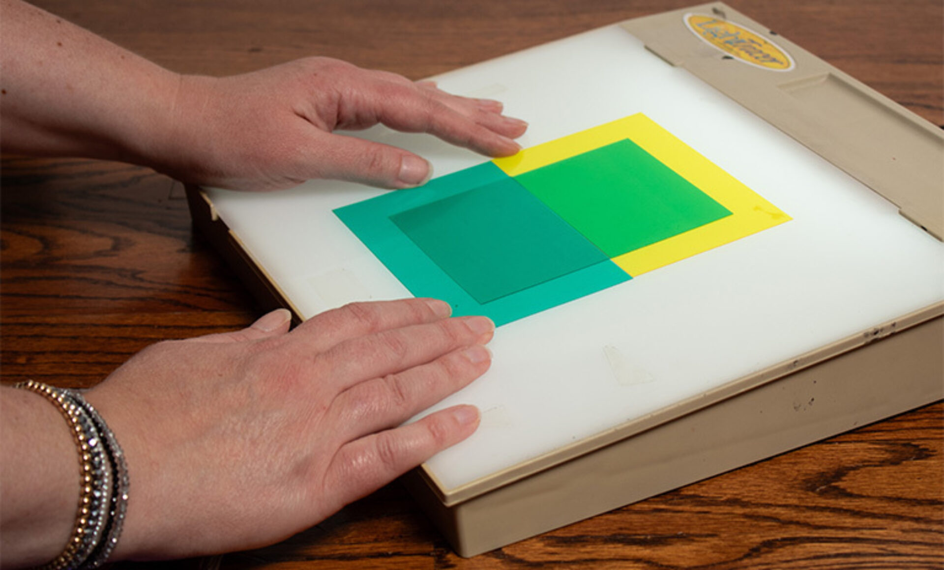 Two hands arranging translucent green and yellow square overlays on a white light box, creating a darker teal overlap, on a wood table.