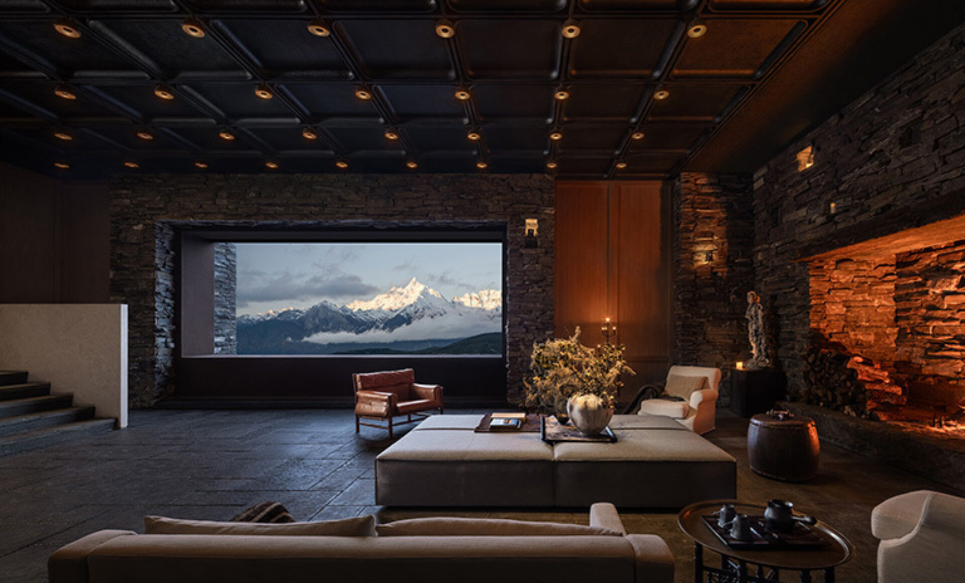 Warm stone lounge with low sofas facing a panoramic window framing snow-capped peaks above clouds, lit by a glowing fireplace.