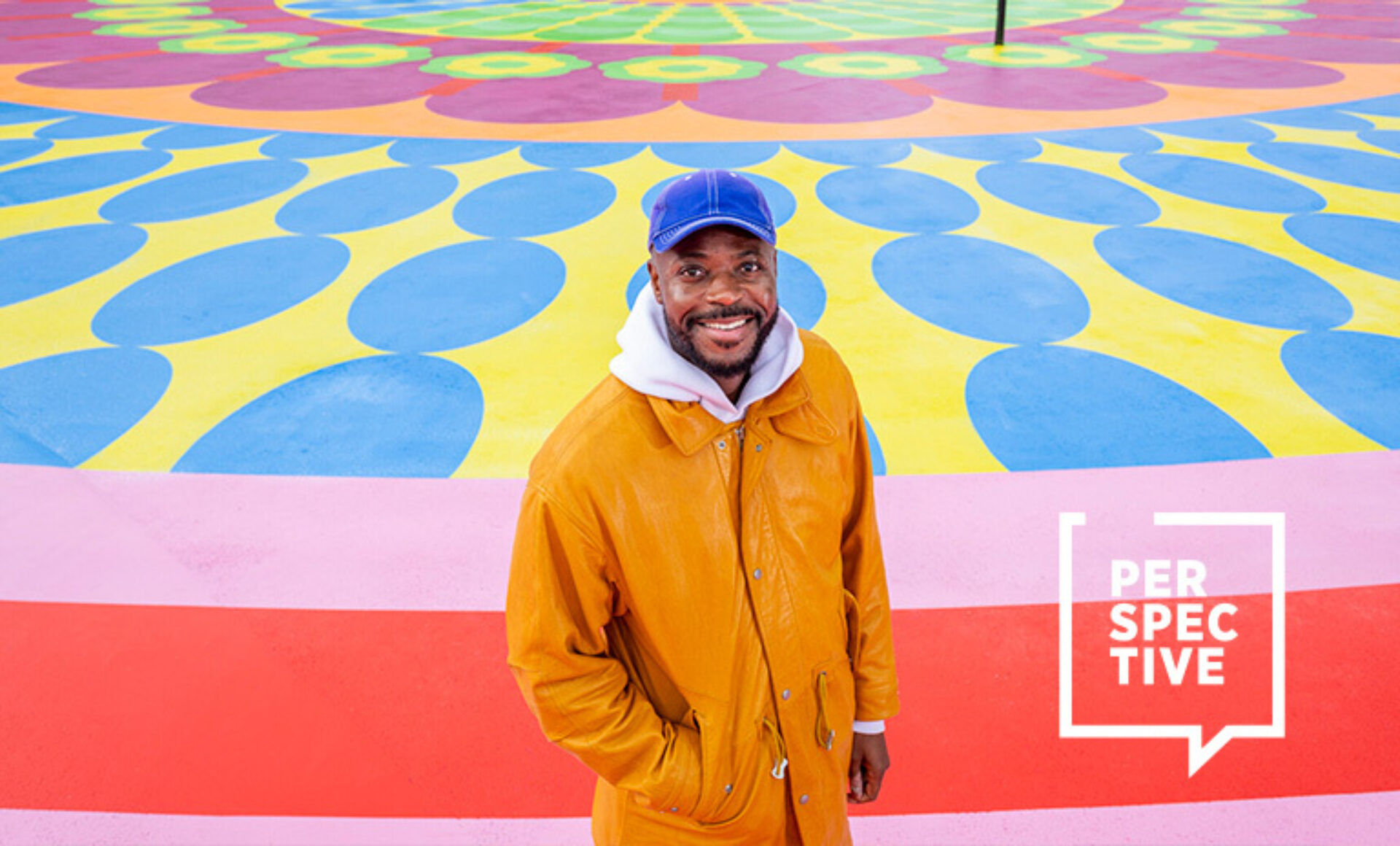 Smiling man in an orange coat and blue cap stands before a colorful circular mural with concentric petal patterns and bold stripes.