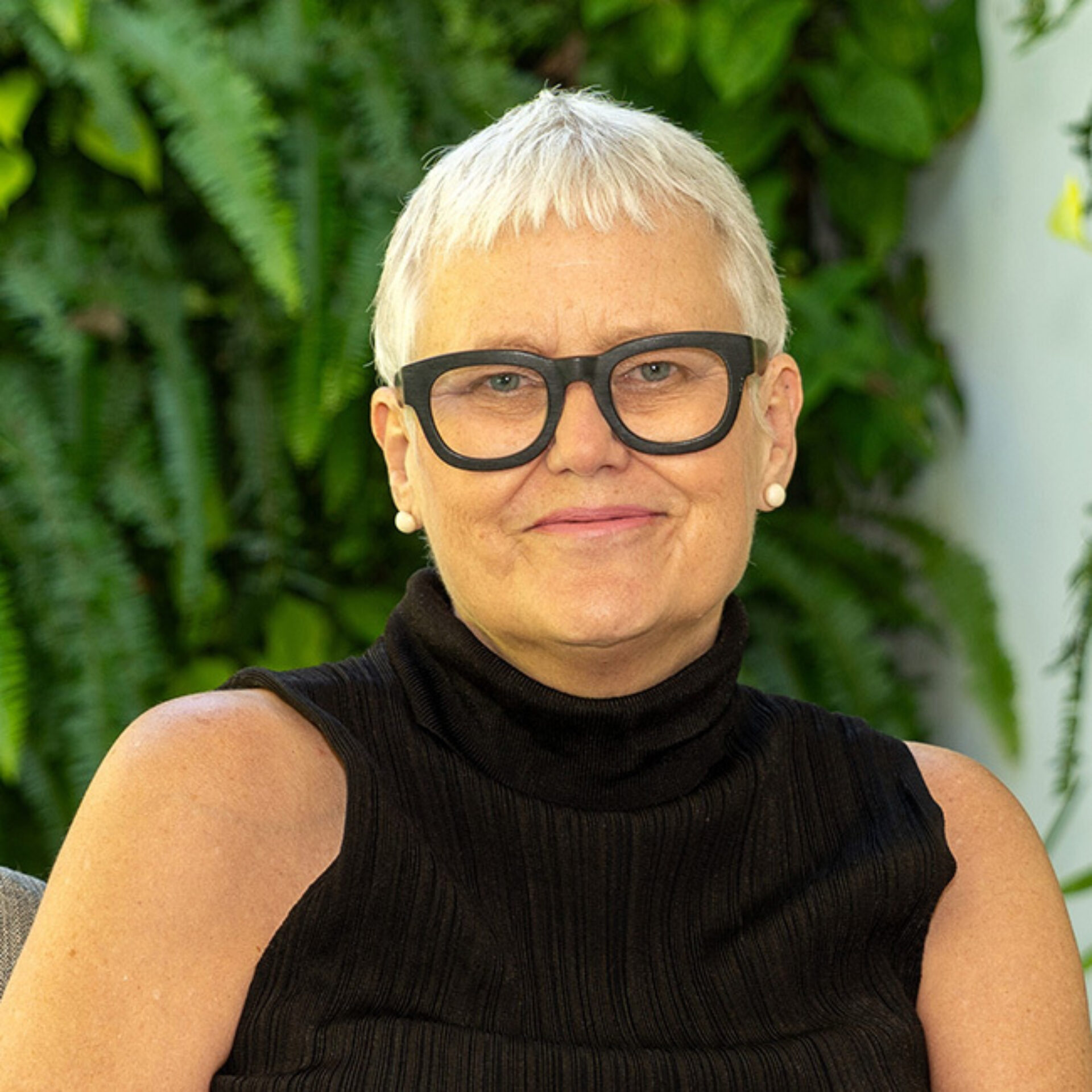 Smiling older person with cropped white hair, bold black-frame glasses and pearl studs, wearing a sleeveless black turtleneck against ferns.