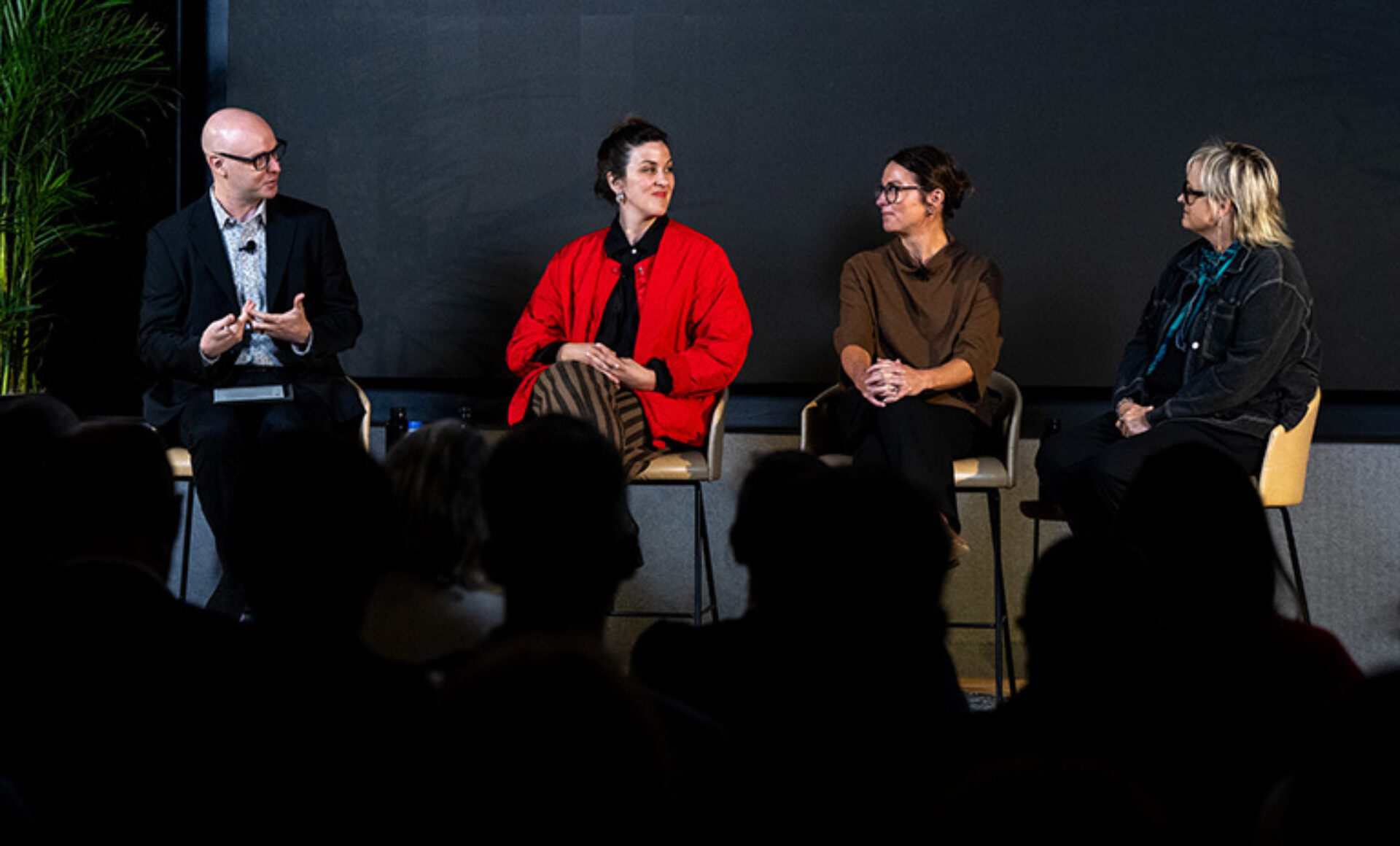 Four panelists on stage with dark backdrop: a bald man gestures, a woman in a red jacket watches, two women listen, audience silhouettes.