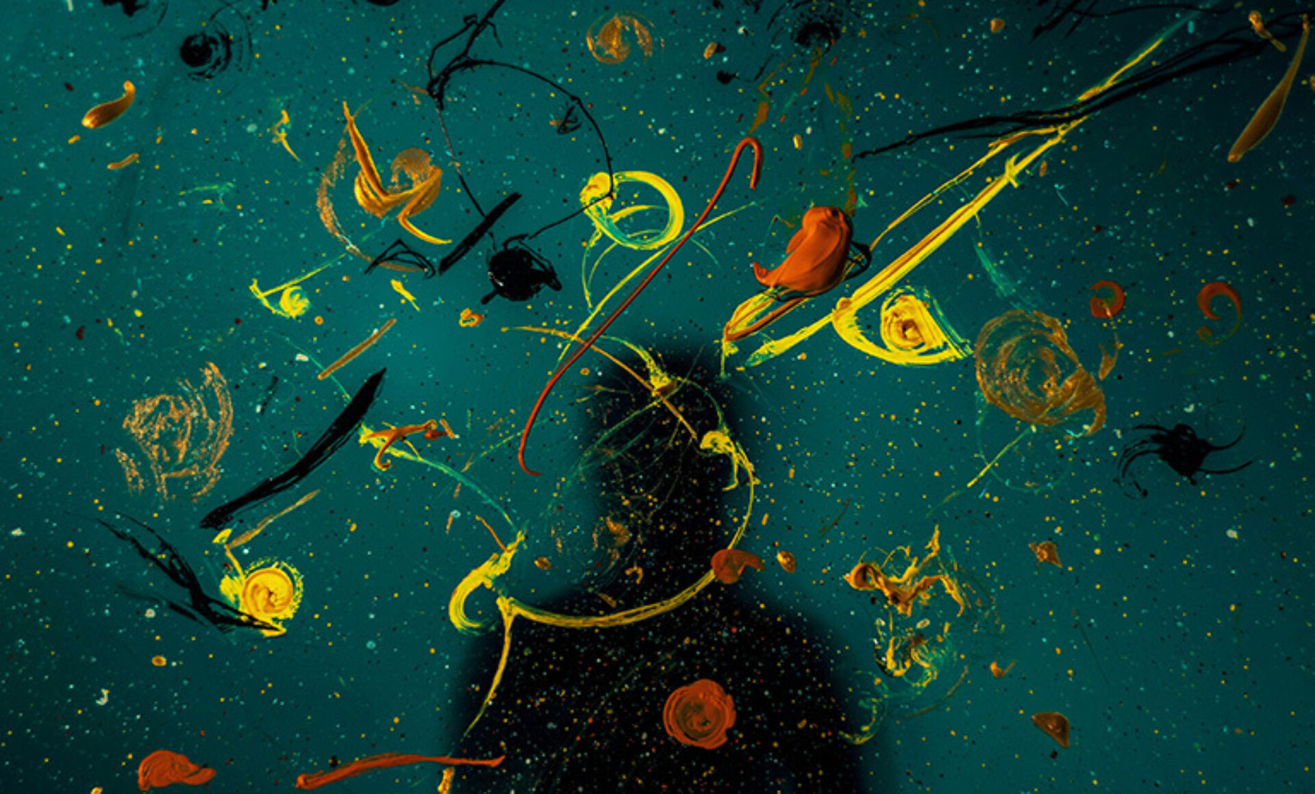 A dark surface displays vibrant swirls of yellow, orange, and green paint, creating an abstract design, with a faint silhouette in the background.
