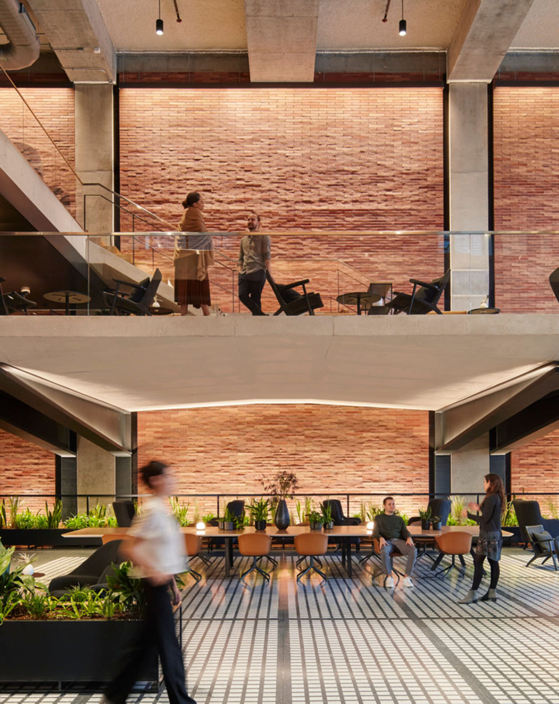 A modern indoor space with two levels, featuring a warm brick wall, seating areas, and people interacting amidst greenery and stylish decor.